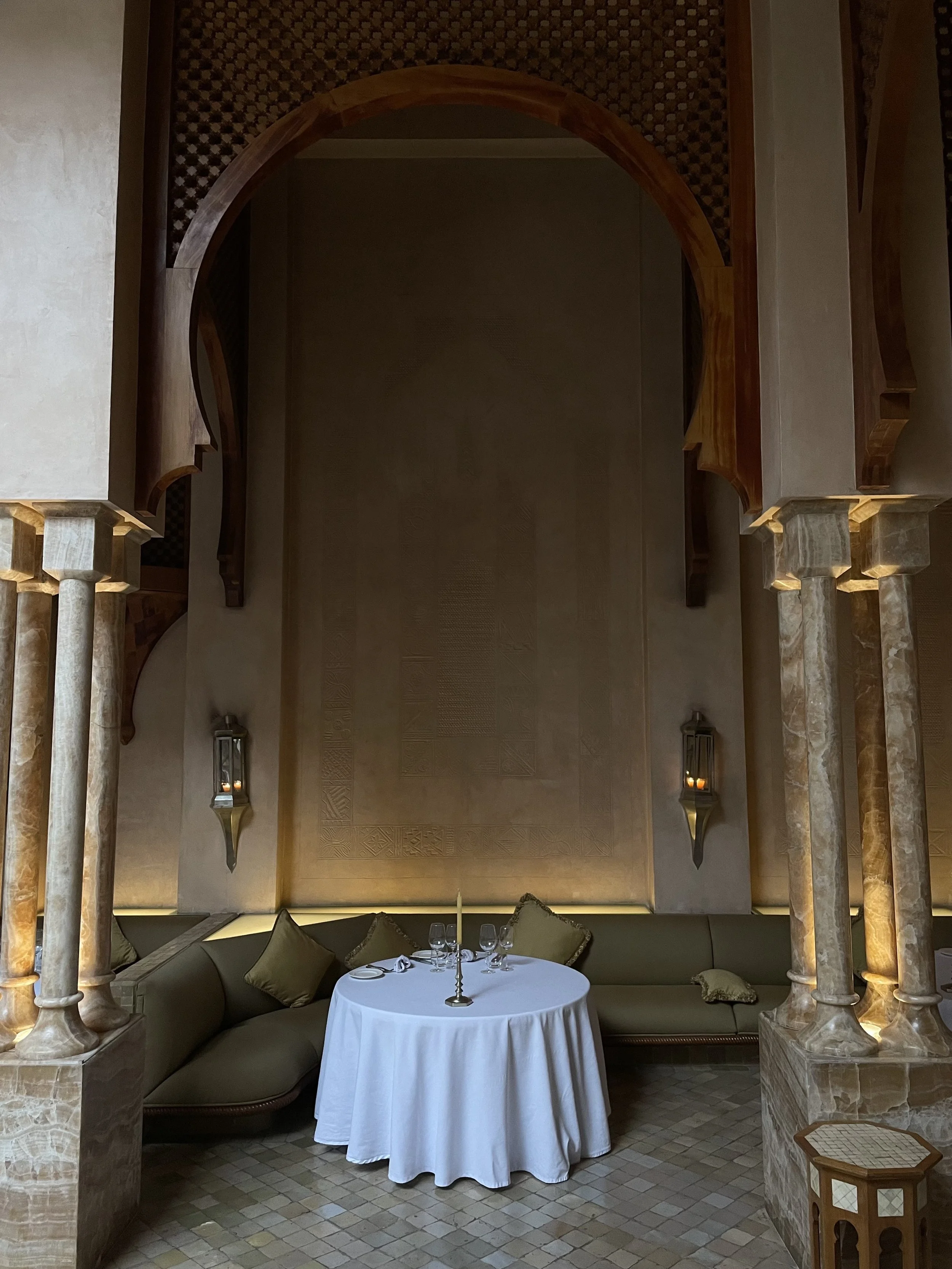 Elegant dining area with a round table covered with a white cloth, set with glasses and cutlery, surrounded by cushioned seating, ornate arches, and ambient lighting.