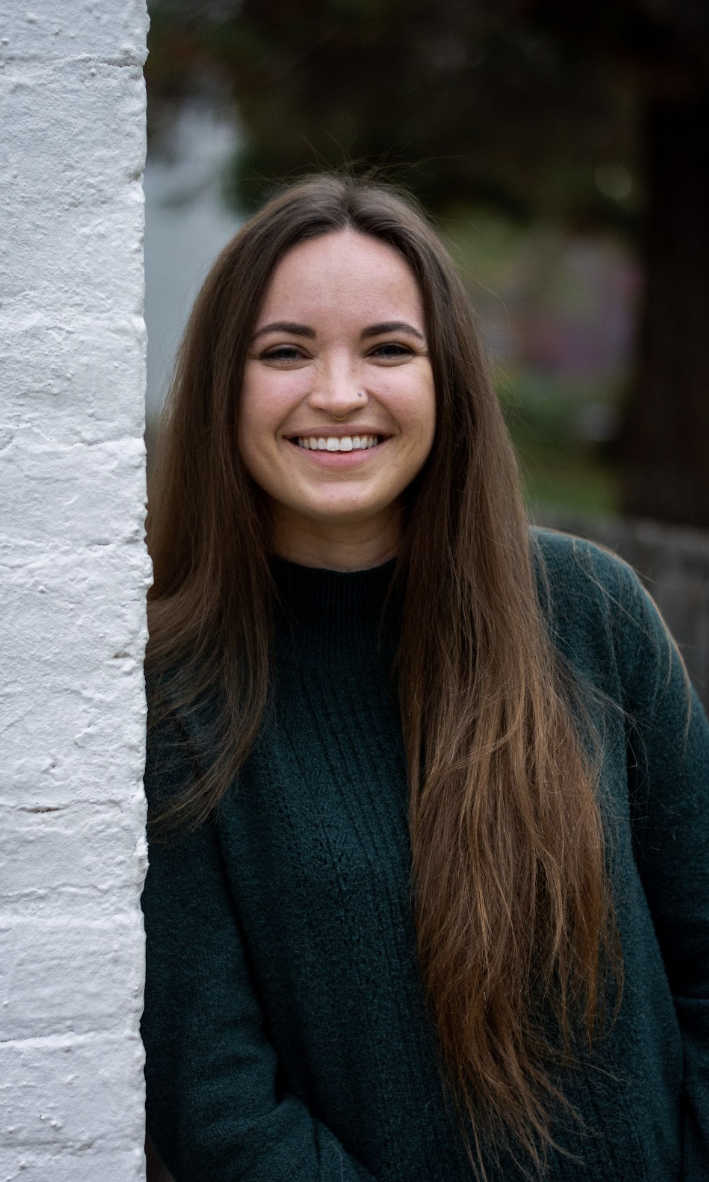 A young female speech-language pathologist with long brown hair, smiling, standing outdoors next to a white brick wall, wearing a dark green sweater.