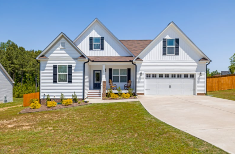 White two-story house with black shutters, front porch with chairs, attached garage, and a well-maintained lawn under a clear blue sky.