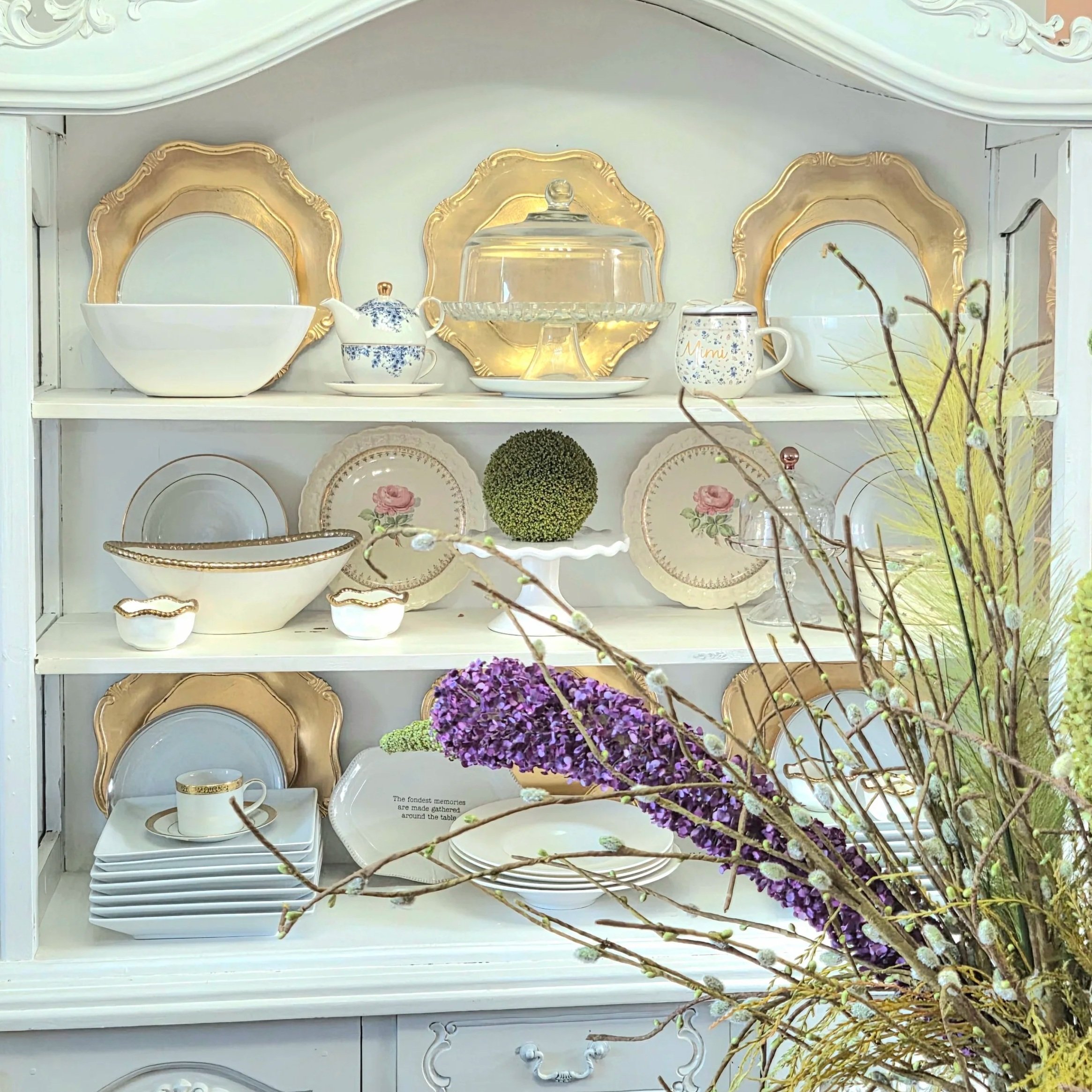 White china cabinet displaying an assortment of decorative and dishware items including plates, bowls, cups, teapot, and cake stand, with a bouquet of purple, green, and beige flowers in the foreground.
