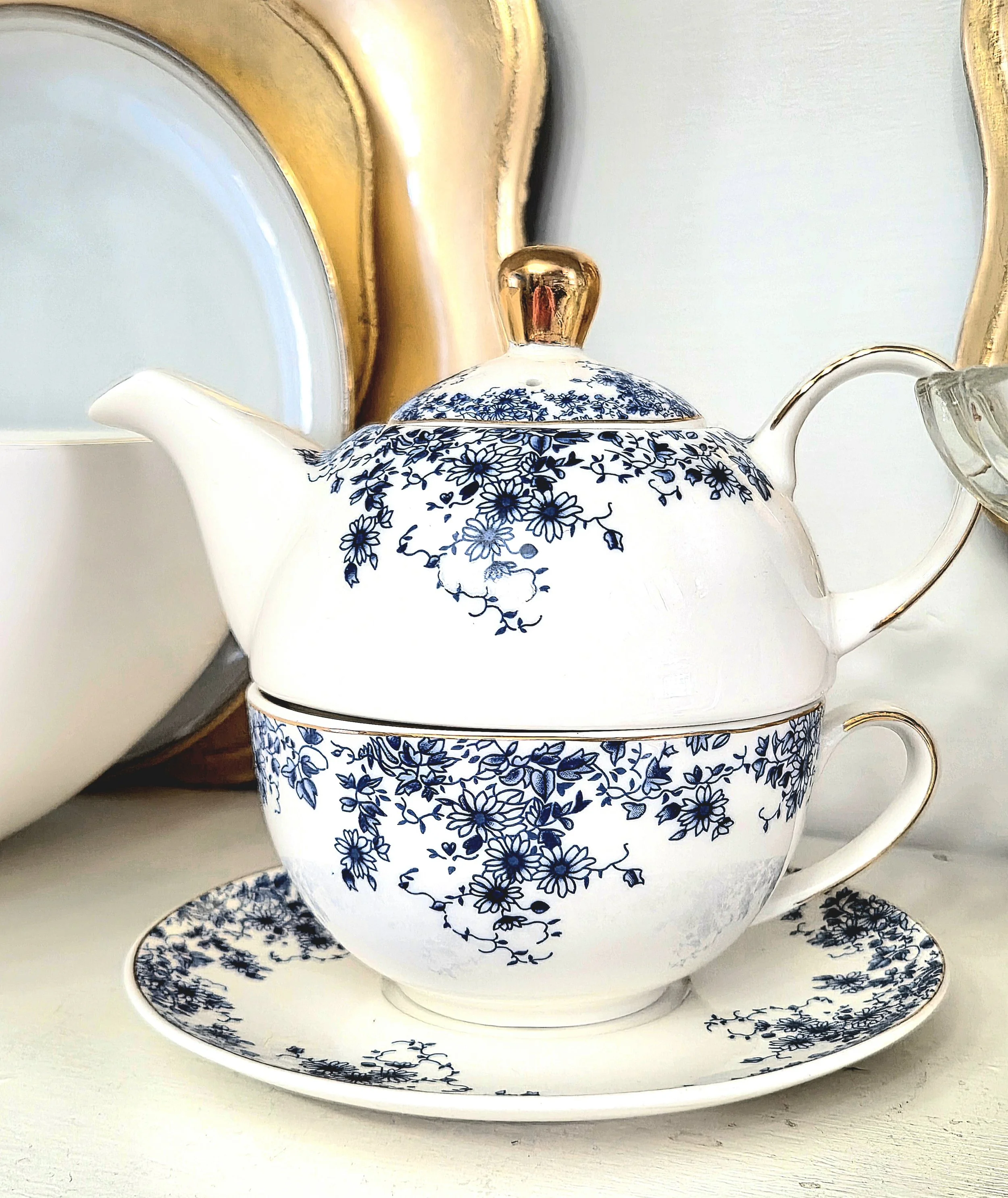 A white teapot with blue floral patterns stacked on a matching teacup and saucer, gold accent lid, in front of gold-framed mirrors.
