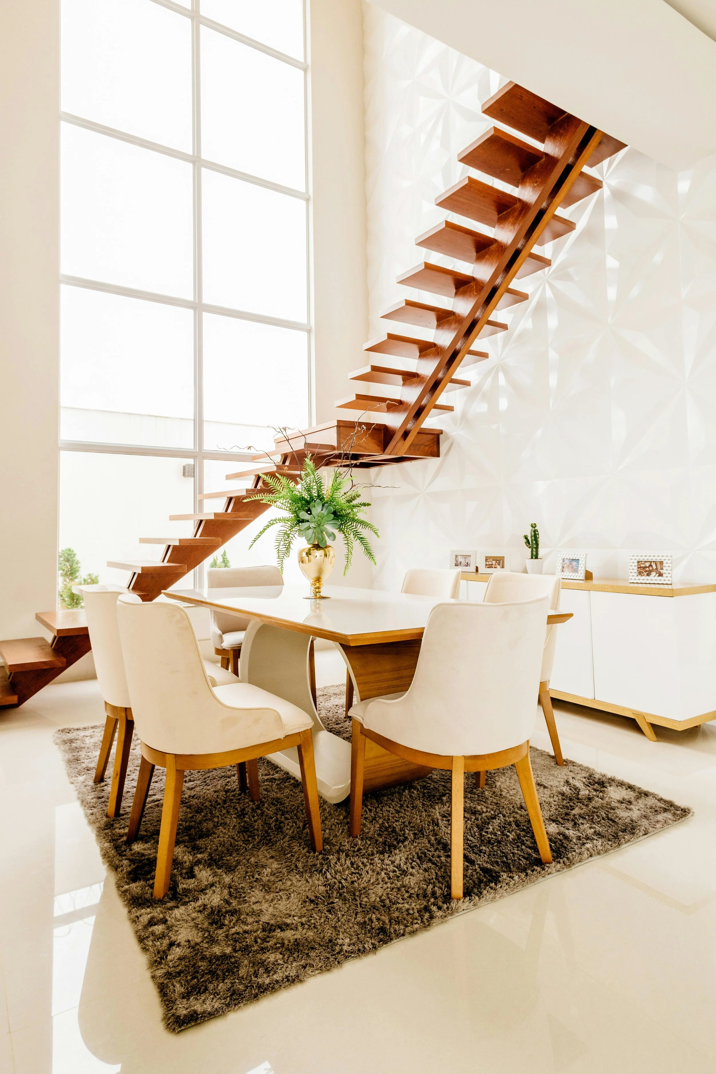 Modern dining area with a wooden table, six cream upholstered chairs, a shaggy gray area rug, a large window, a white textured wall, a green plant in a gold vase, and a white sideboard with framed photos and a small cactus.