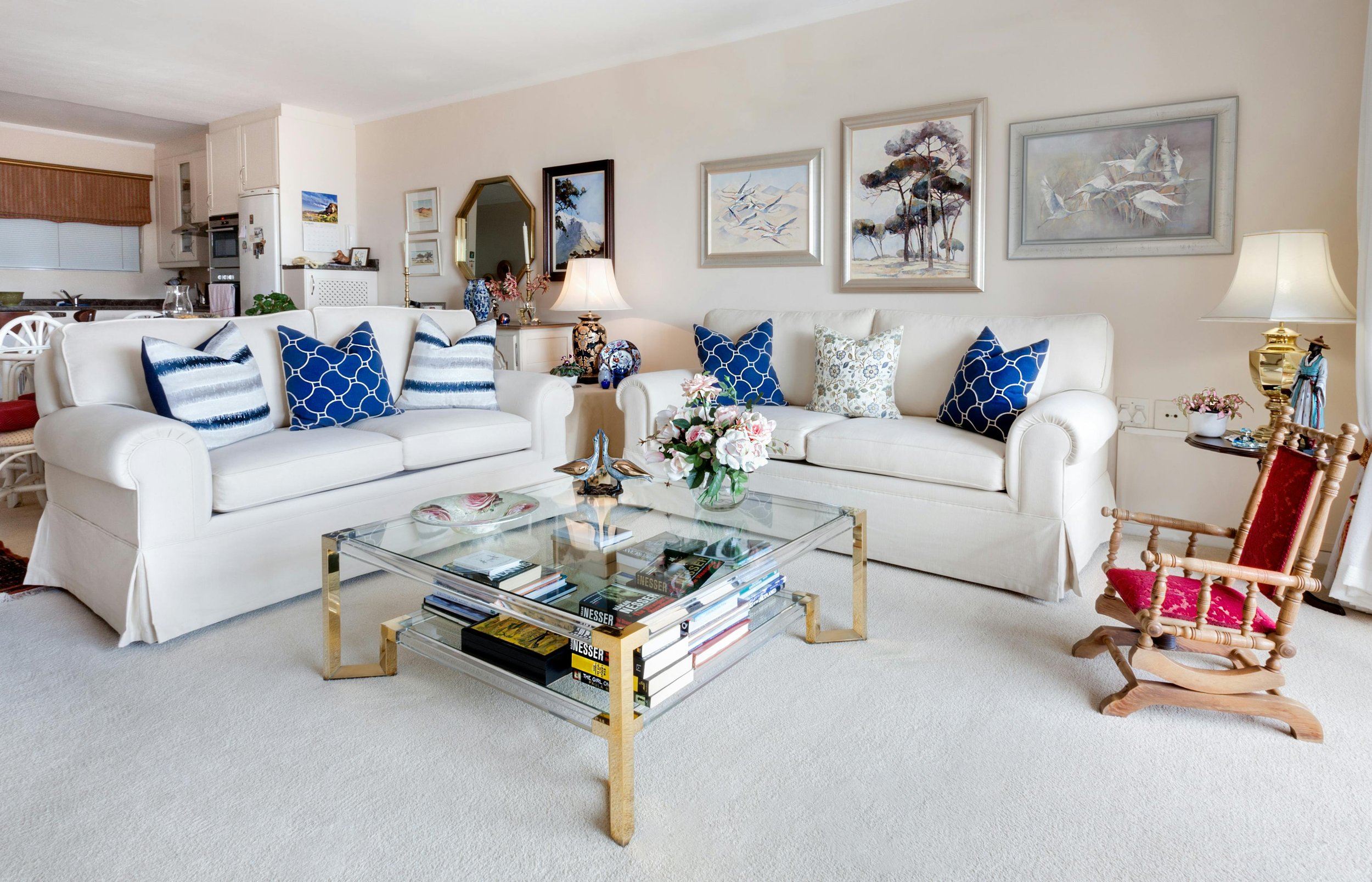 Living room with white sofas, blue and white throw pillows, a glass coffee table with books, a bouquet of flowers, and wall art, with a wooden rocking chair with red cushions nearby.