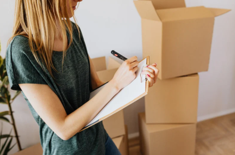 Person writing on a clipboard with moving boxes in the background.