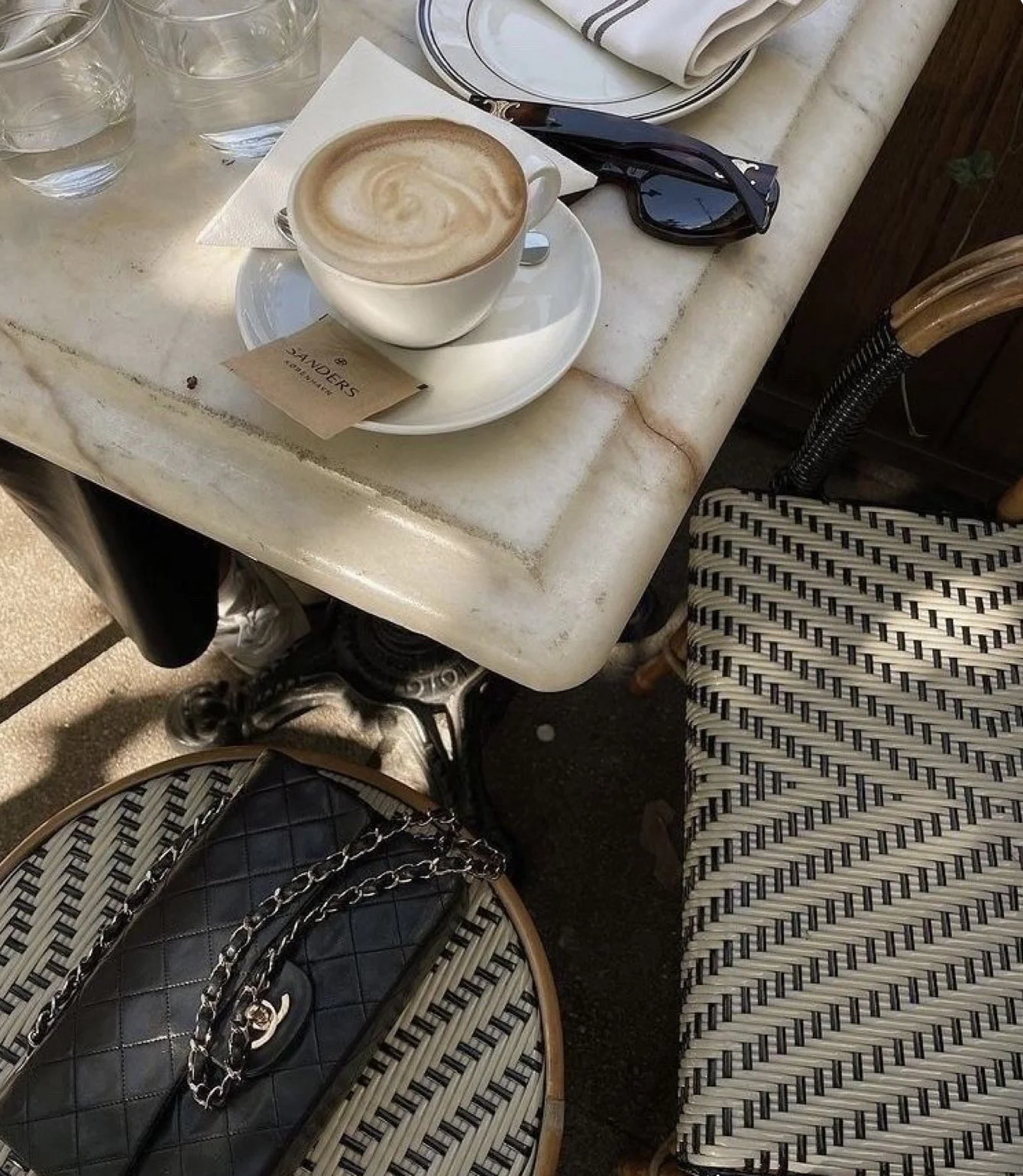 A marble table with a cup of cappuccino, three glasses of water, a pair of sunglasses, a napkin, and a stack of plates. A black quilted handbag with a chain strap is on a woven chair beside the table.