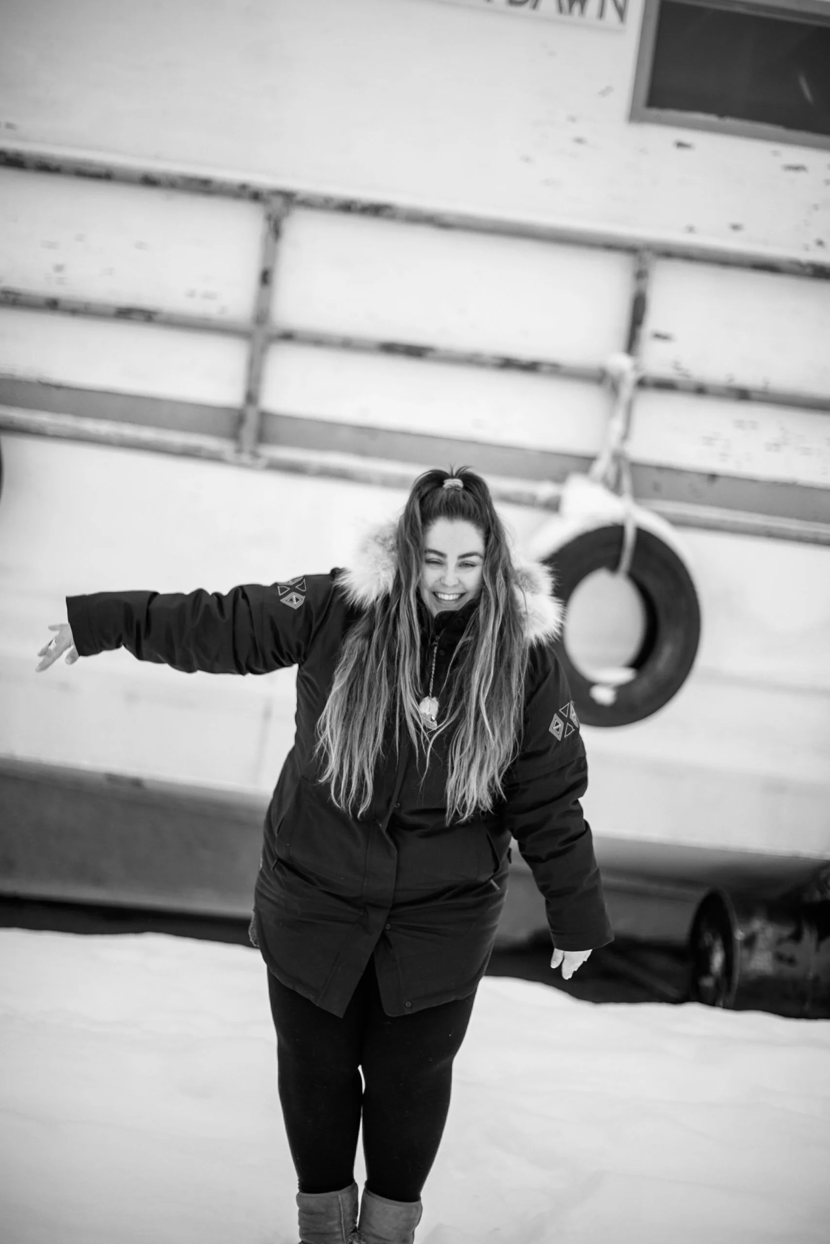 A woman with long hair in a ponytail, wearing a winter coat with fur trim, smiling and waving while standing outdoors in the snow.