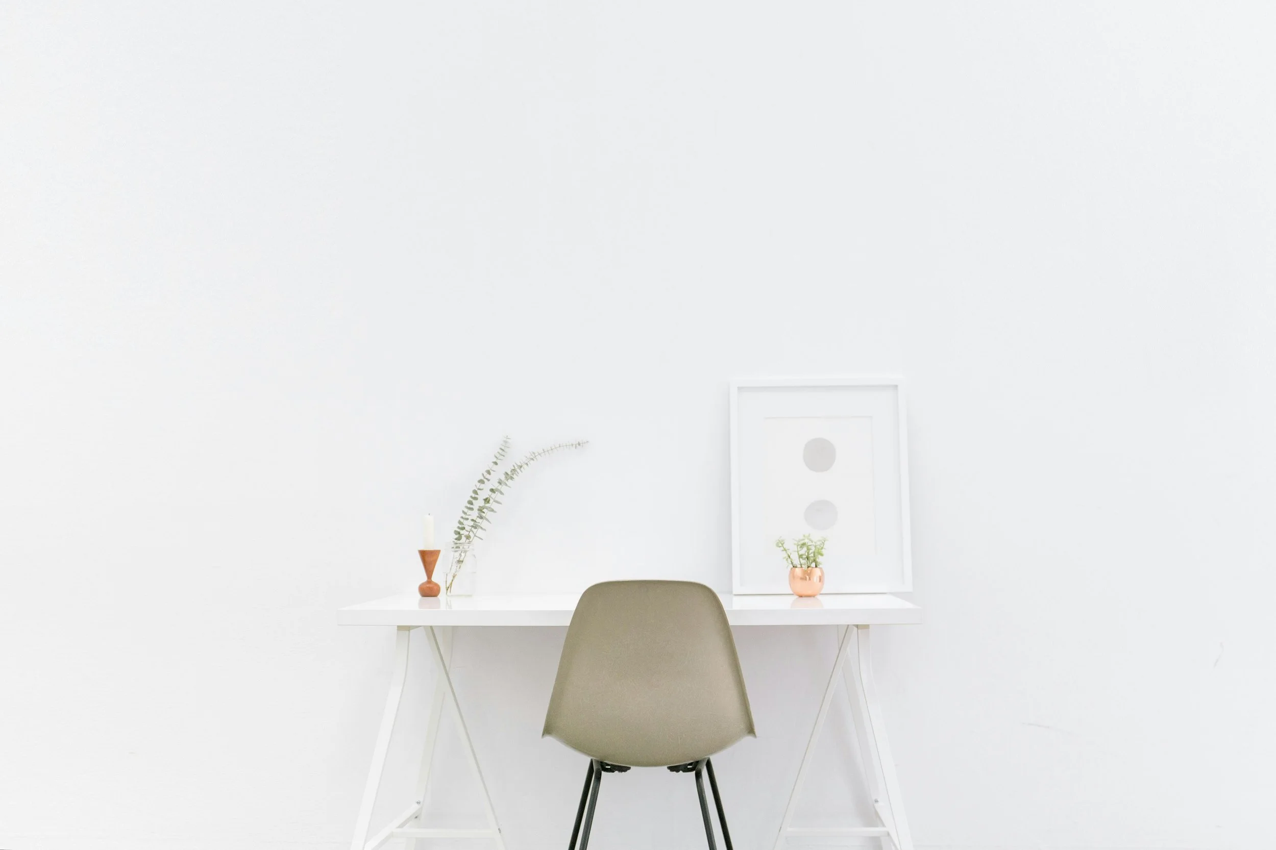 Minimalist white desk with a chair, a framed art piece, small potted plant, candle, and decorative branches against a white wall.