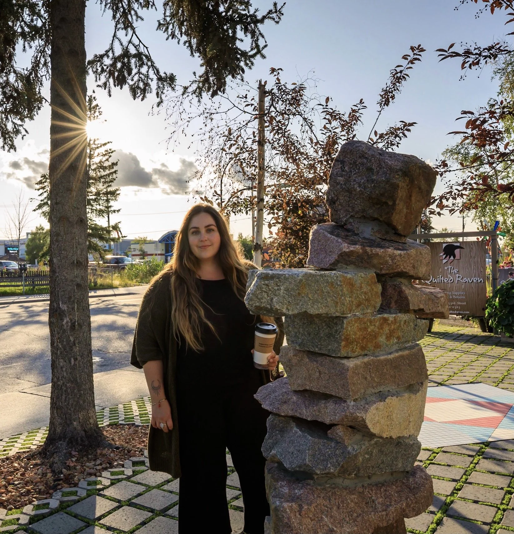 A woman with long brown hair holding a coffee cup, standing outdoors next to a tall rock sculpture, with trees and a sunny sky in the background.