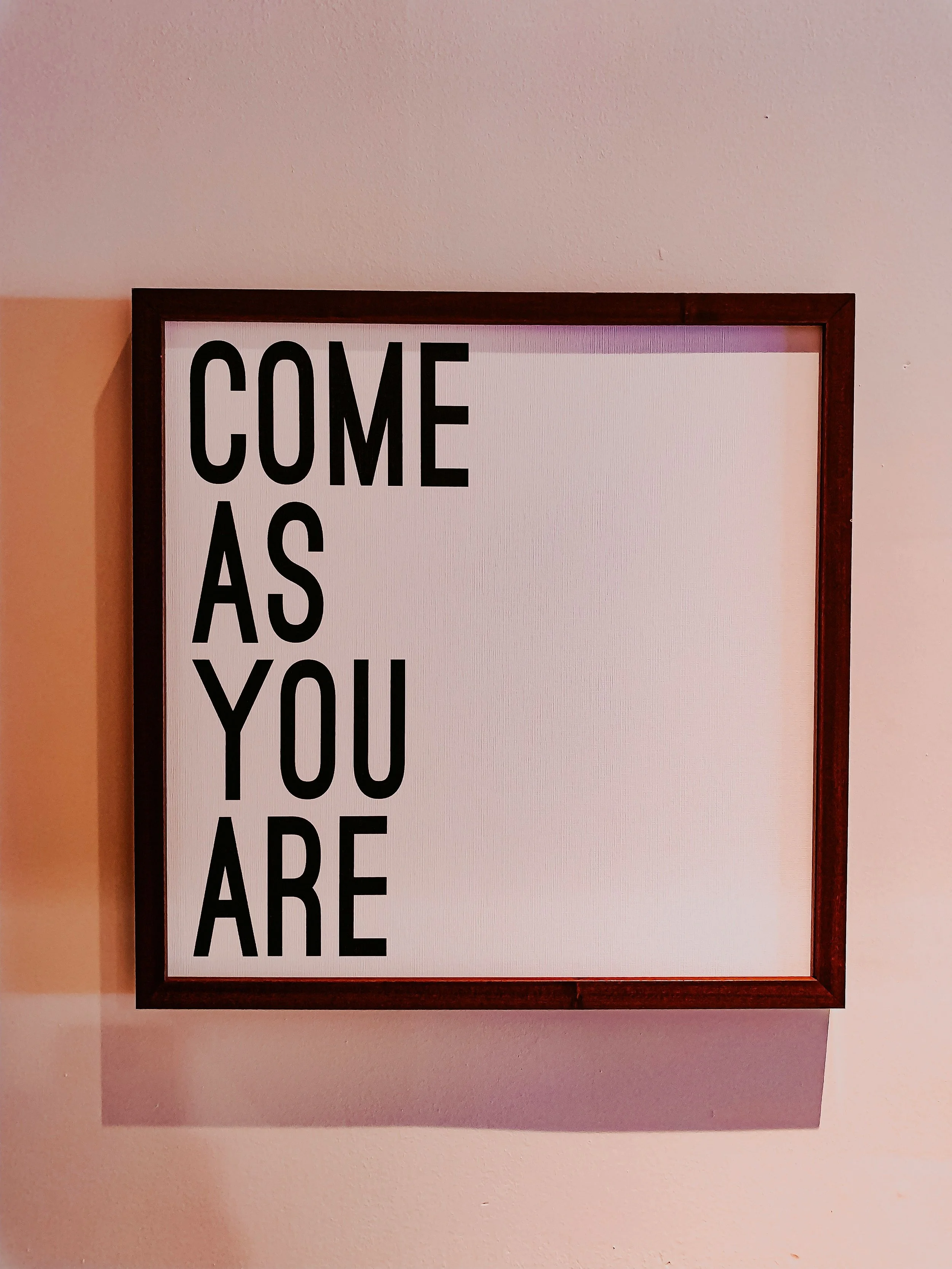 A framed sign on a wall with the text "COME AS YOU ARE" in large black letters.