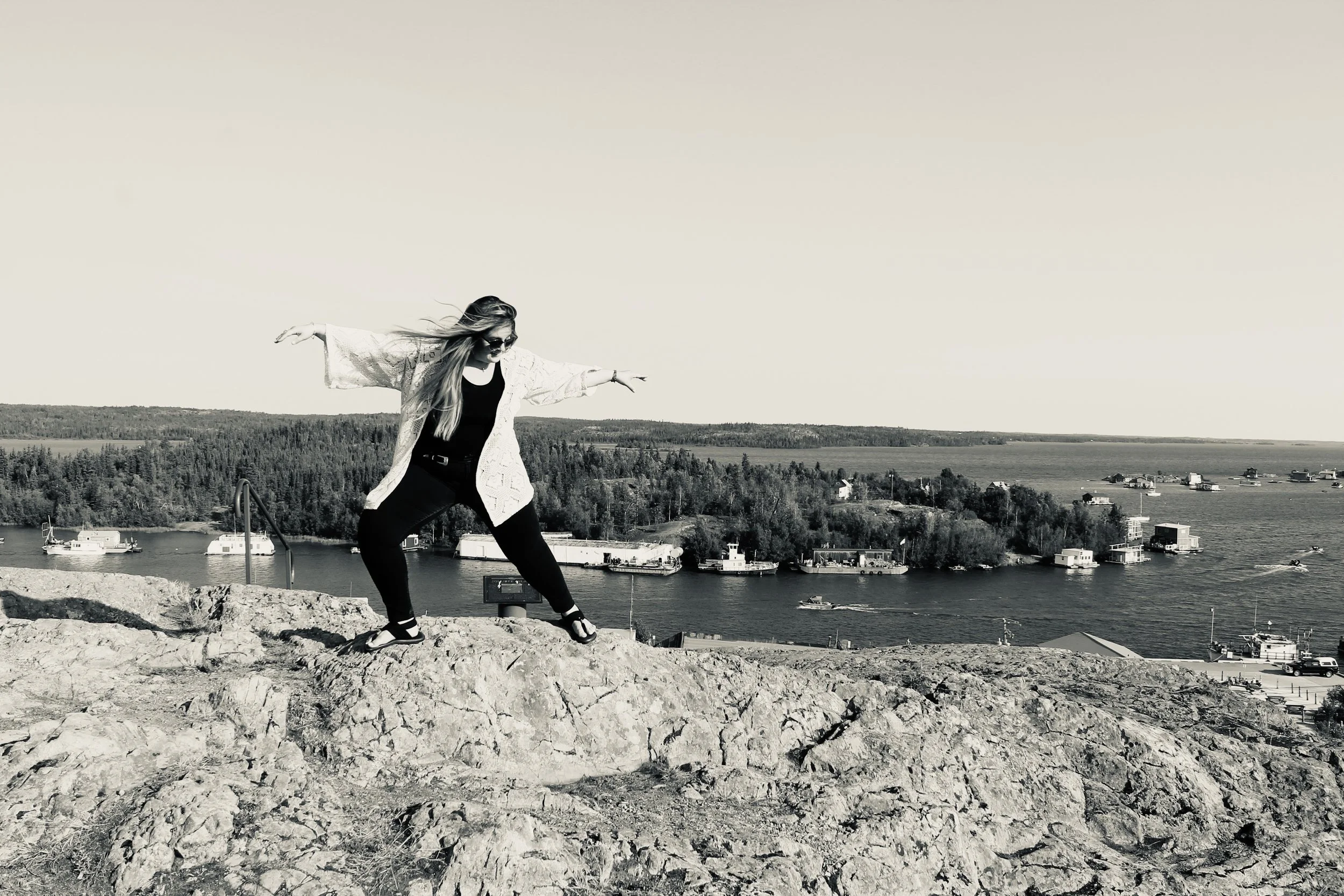 A woman standing on a rocky hilltop with her arms outstretched, overlooking a body of water with boats and houses in the distance, in a black-and-white photograph.
