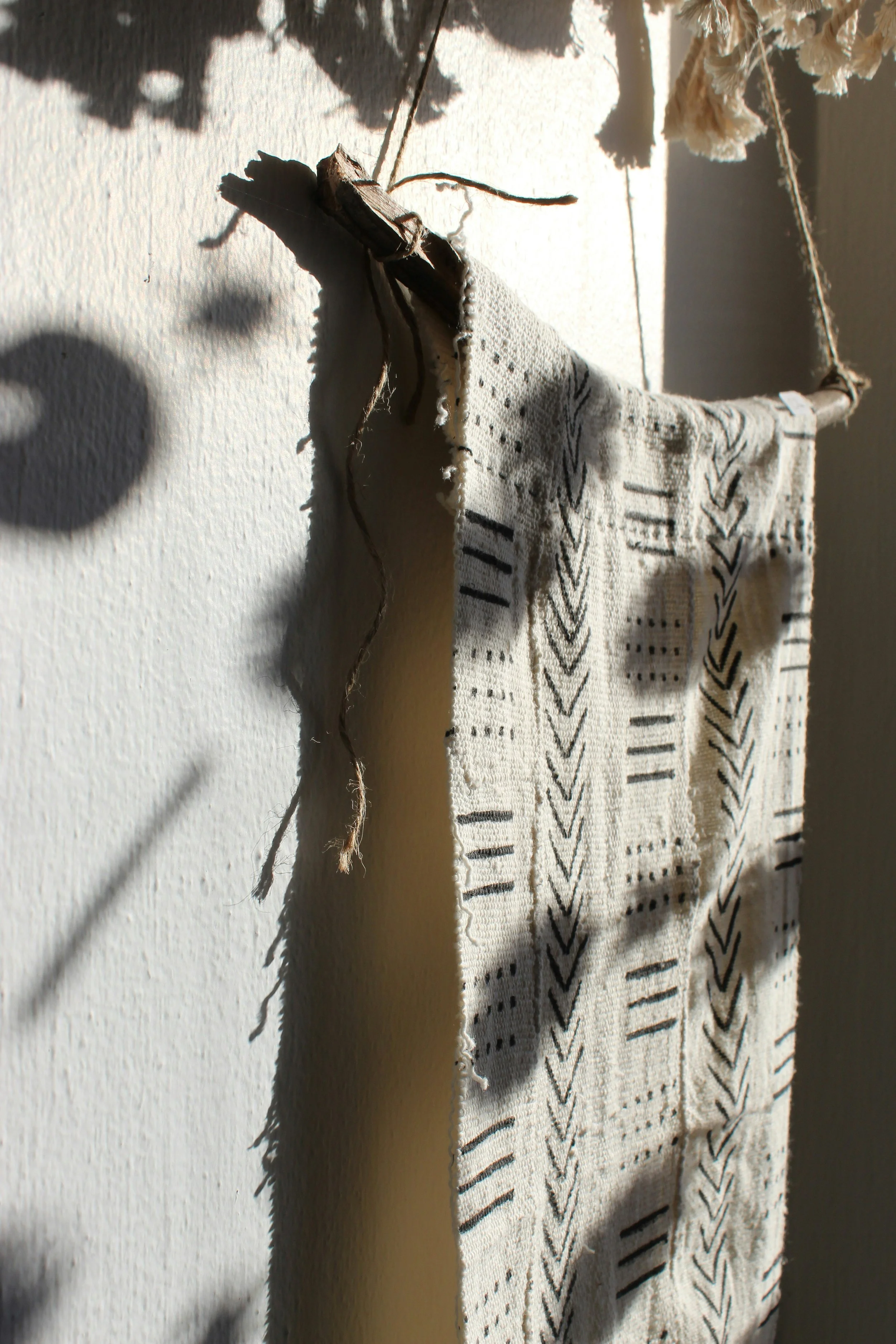 Decorative fabric wall hanging with a wooden stick and string, patterned with black geometric designs, hanging on a wall with shadowed foliage.
