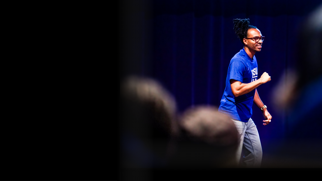 A man on stage giving a presentation, wearing a blue shirt, glasses, and dancing with a smile, with a dark curtain in the background and a blurred audience in the foreground.