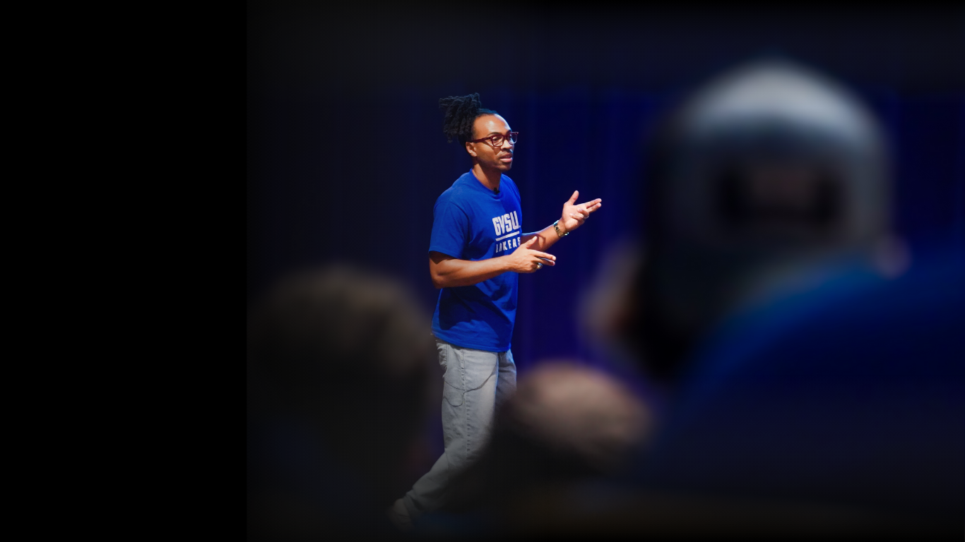 A young man with glasses and dreadlocks wearing a blue shirt and gray pants is speaking on stage with a dark blue curtain backdrop, while an audience watches.