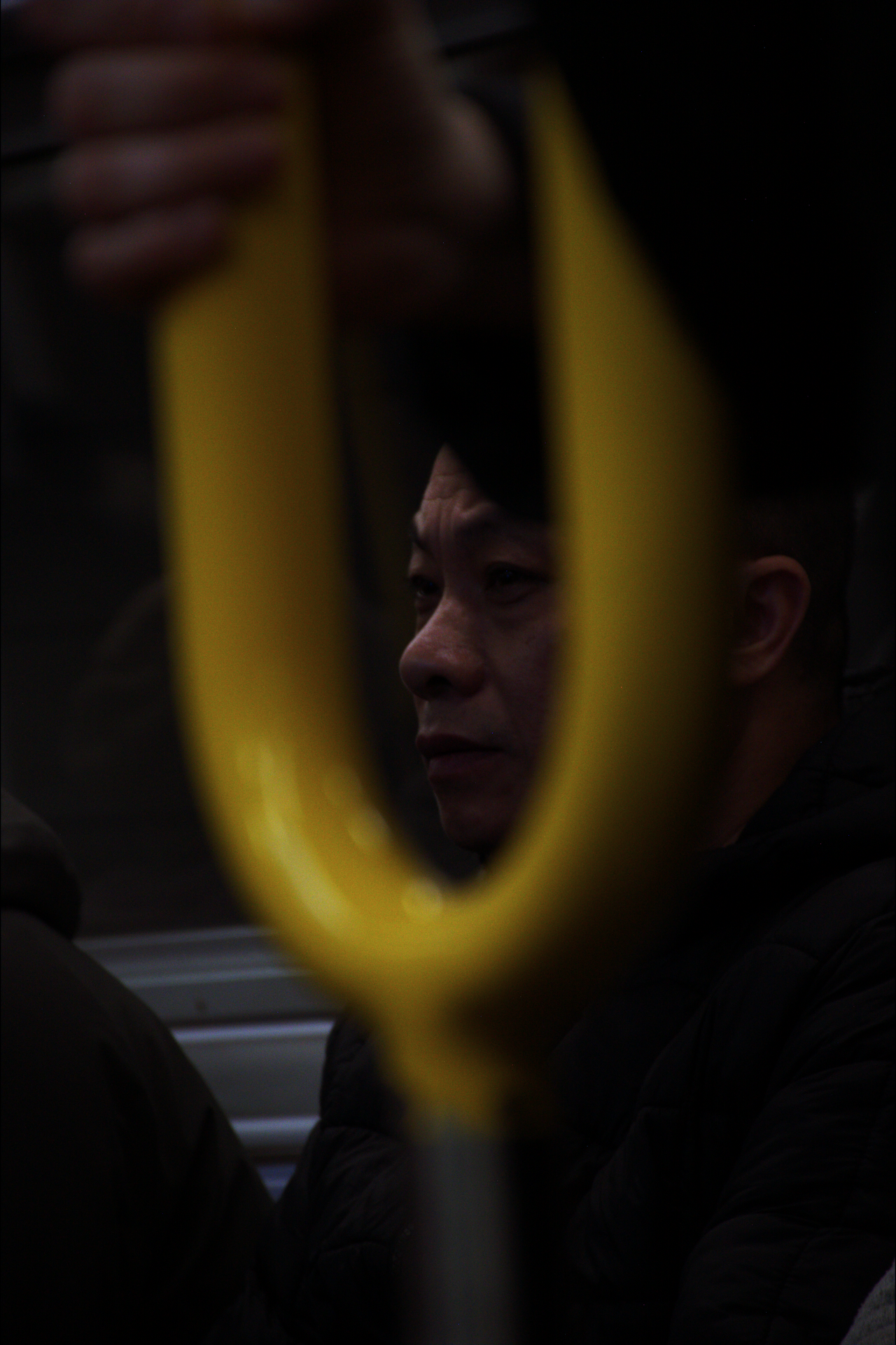 A person's face seen through the reflection of a yellow strap handle on a public transportation vehicle, with a dark background.