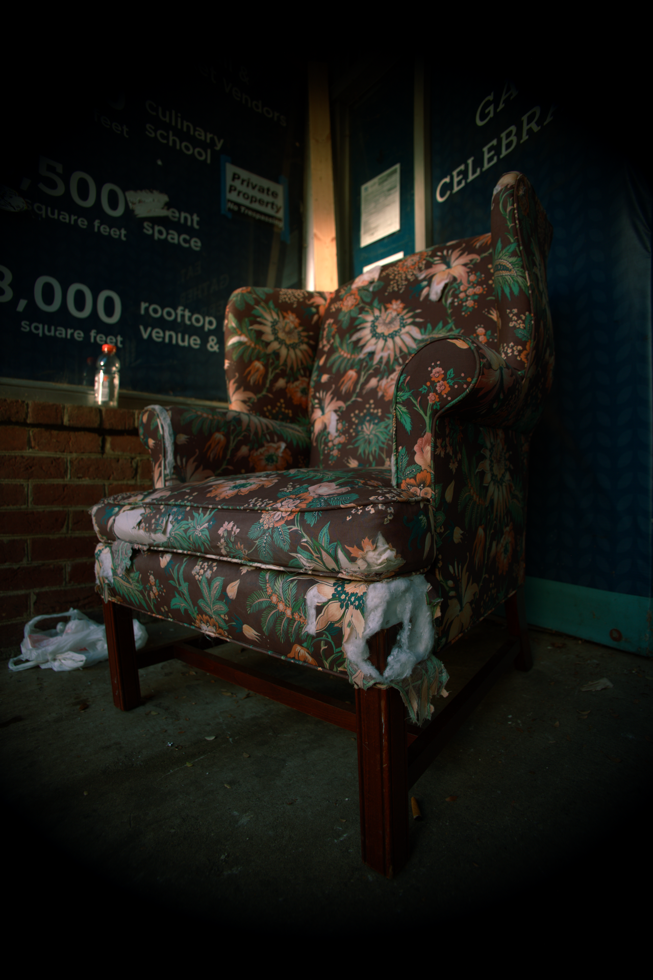 An upholstered armchair with floral fabric, damaged and torn at the front, sitting on a concrete floor in a dimly lit room.