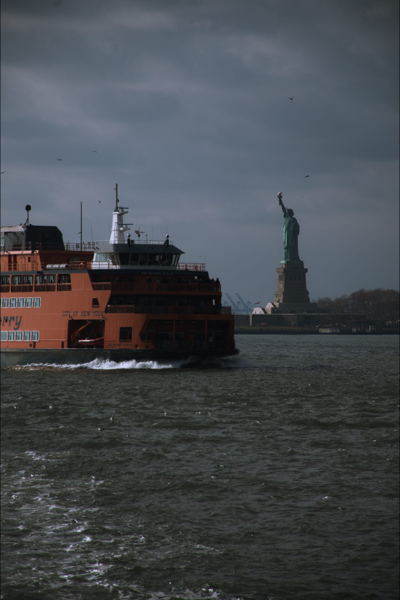 A ferry boat sailing on the water with the Statue of Liberty visible in the background under a cloudy sky.