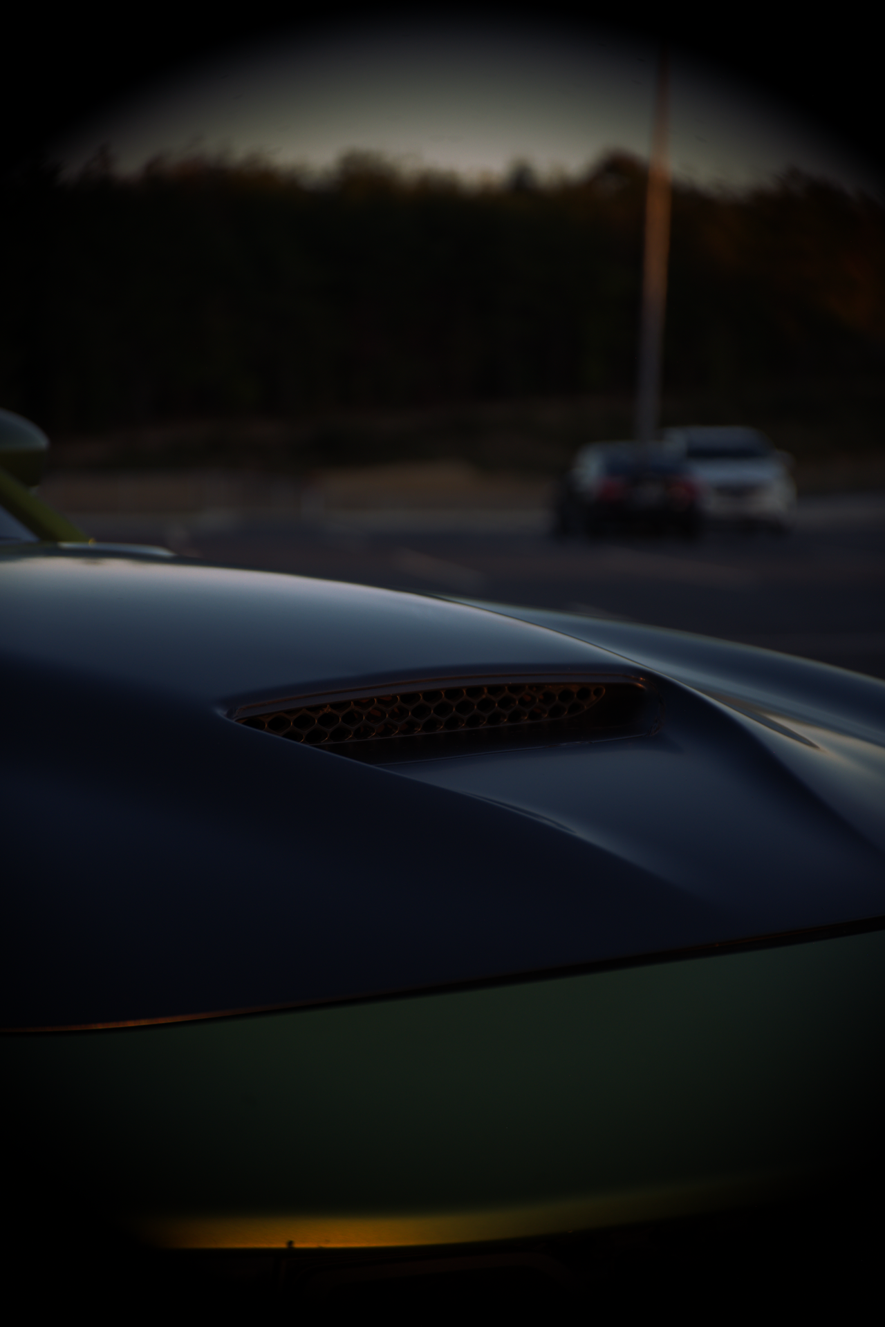 Close-up of a car hood with a vent, blurred background of parked cars and trees, taken during sunset or dusk.