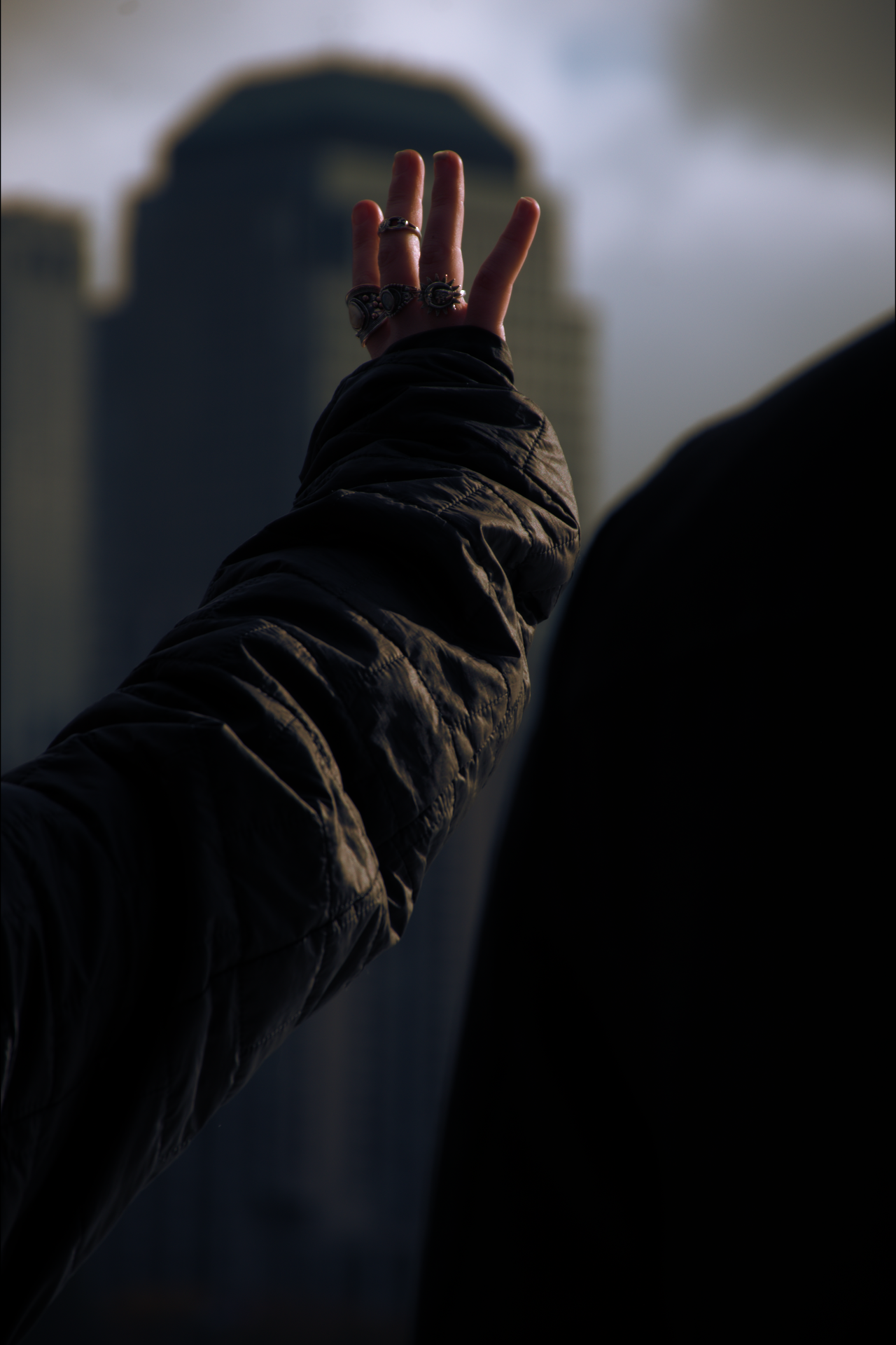 A person wearing a black jacket and multiple rings on their fingers raises their hand in front of a tall building with a distinctive top, under a cloudy sky.