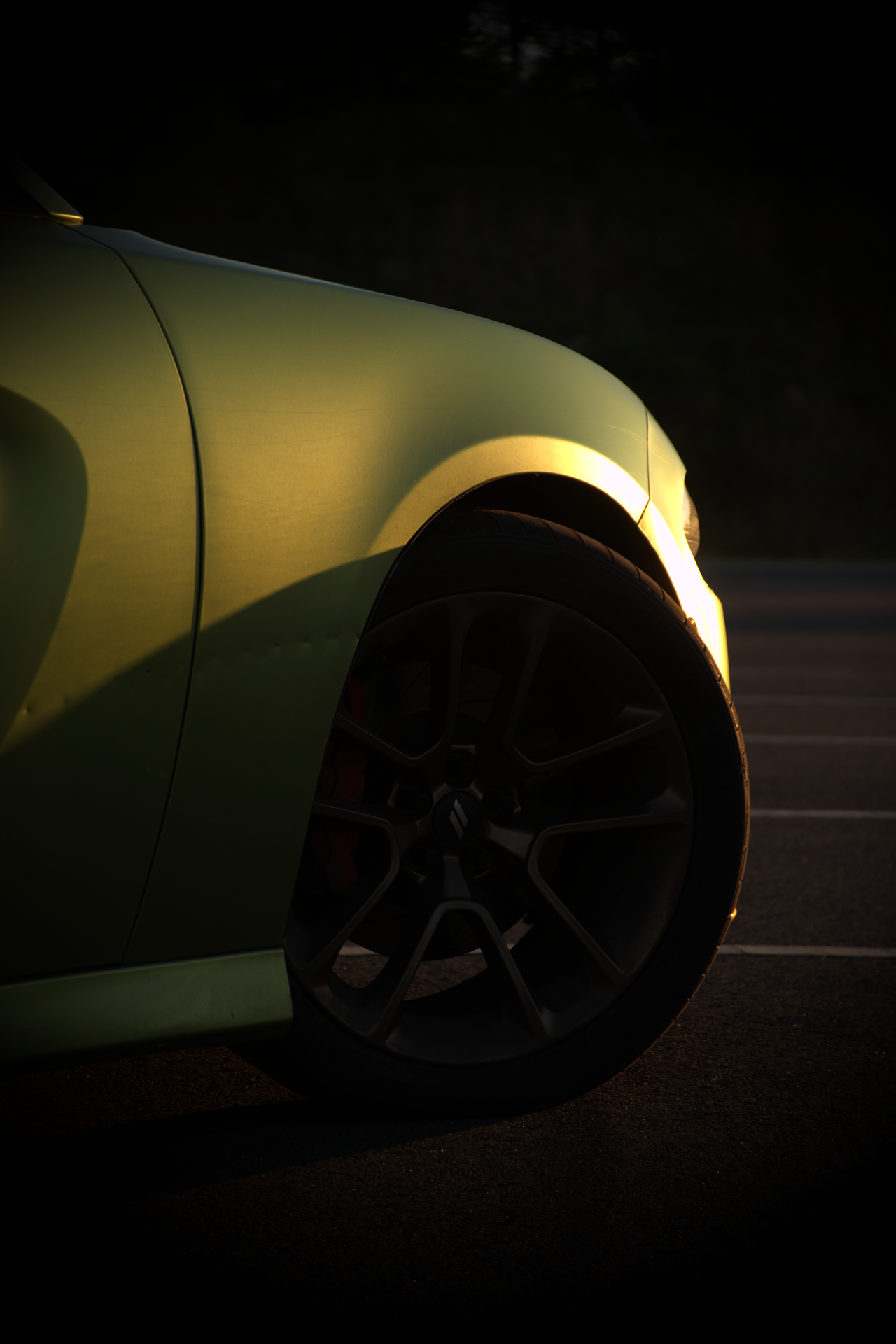 Close-up of the front part of a green sports car, focusing on the front wheel and fender, illuminated by warm sunlight during dusk or dawn with a dark background.