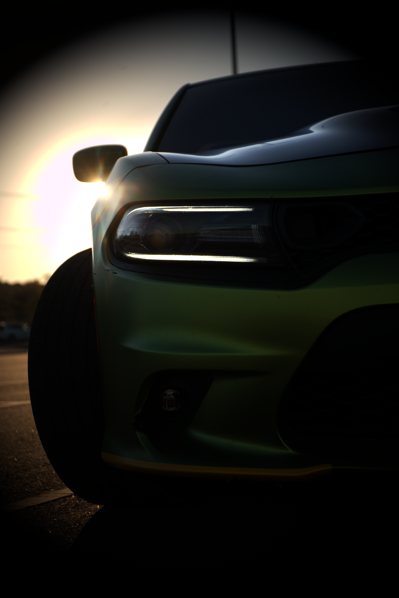 Close-up of the front part of a black car with illuminated headlights, shot at sunset with a blurred background.