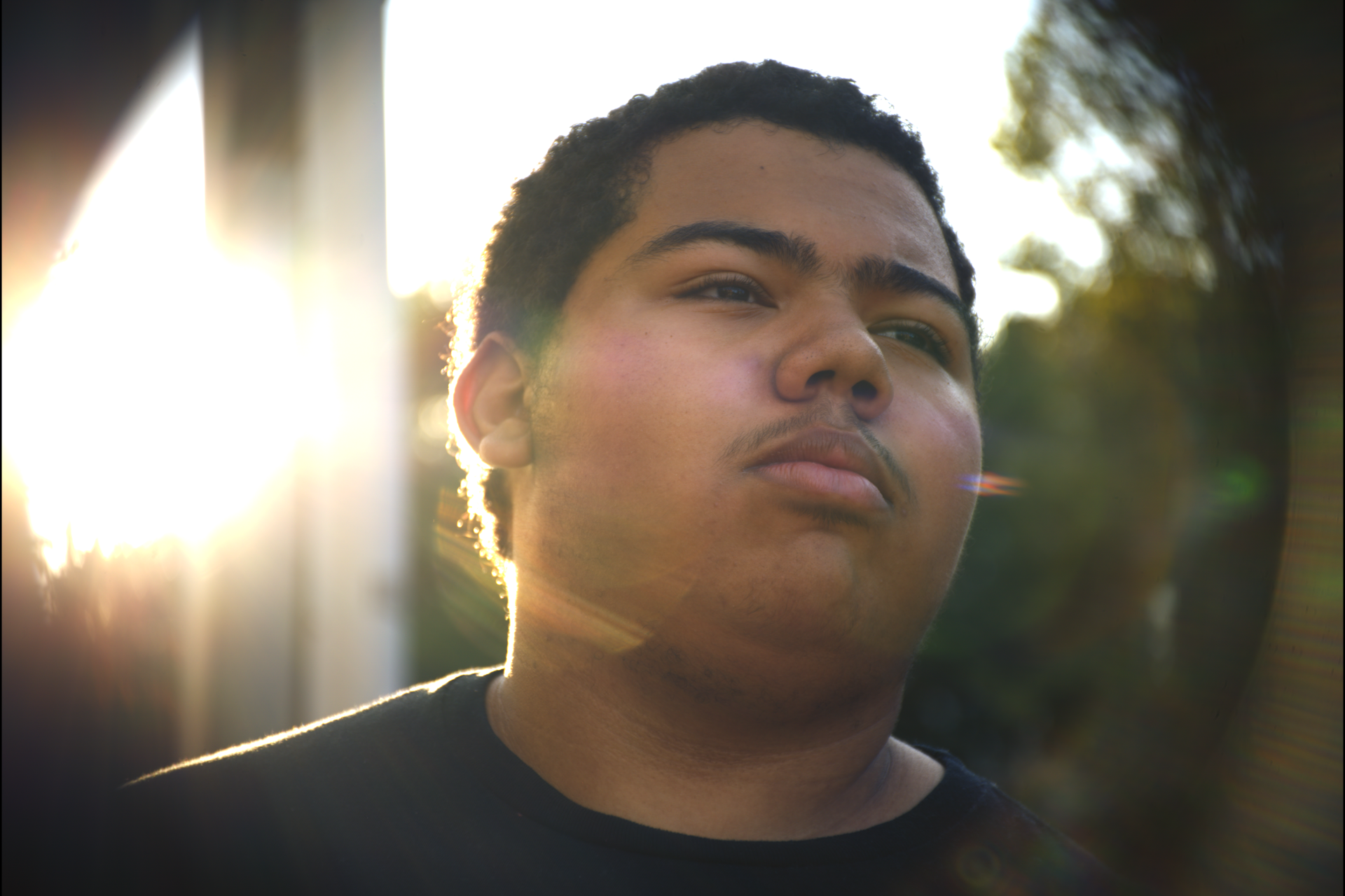 A close-up of a young man with dark hair and a slight mustache, looking to the side with the sun setting behind him, creating a bright backlight.