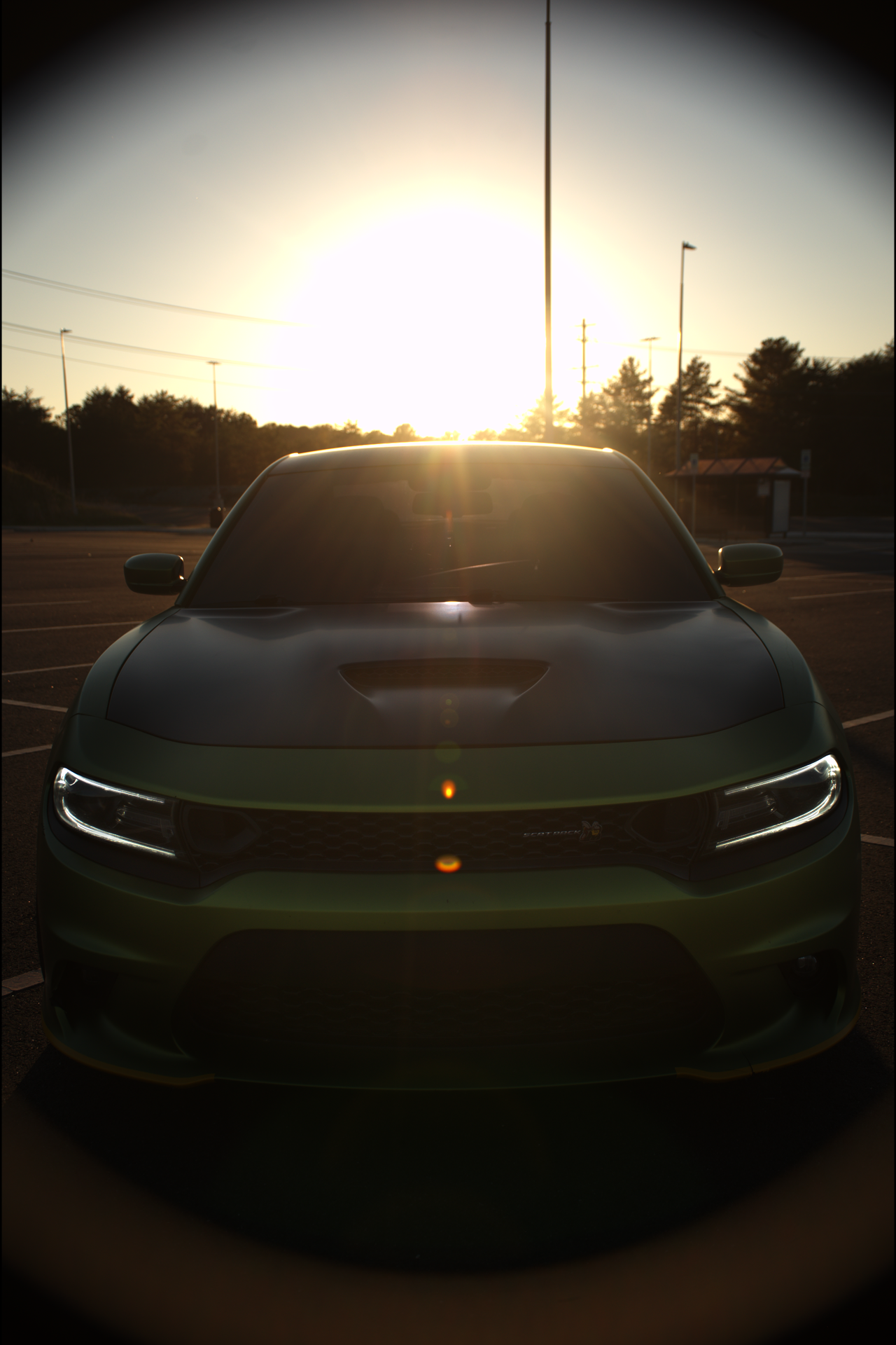Front view of a green Dodge Charger car parked in an empty lot during sunset, with the sun shining behind it, creating a silhouette effect.