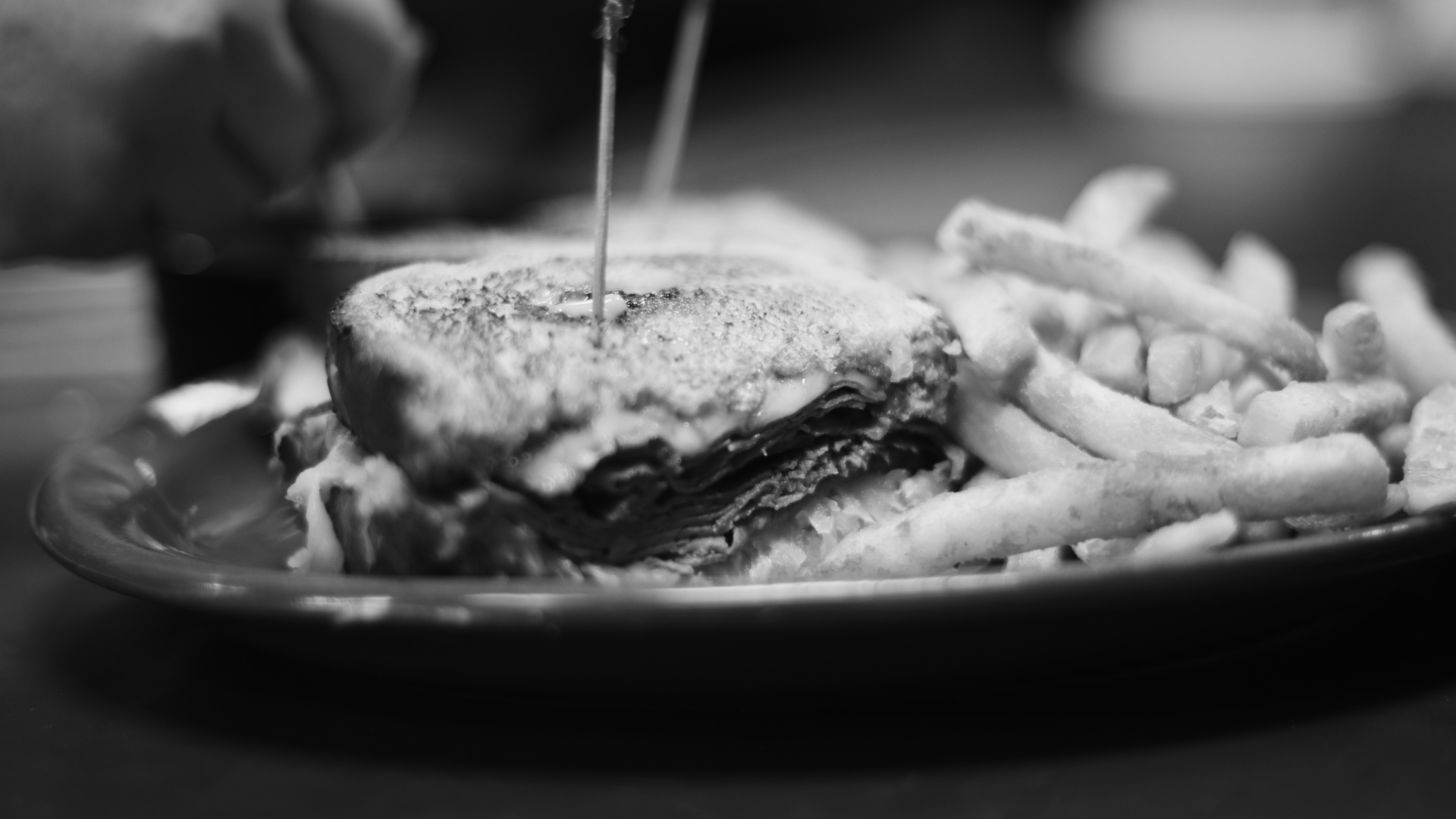 Black and white photo of a plate with a layered burger topped with cheese and fries on the side.