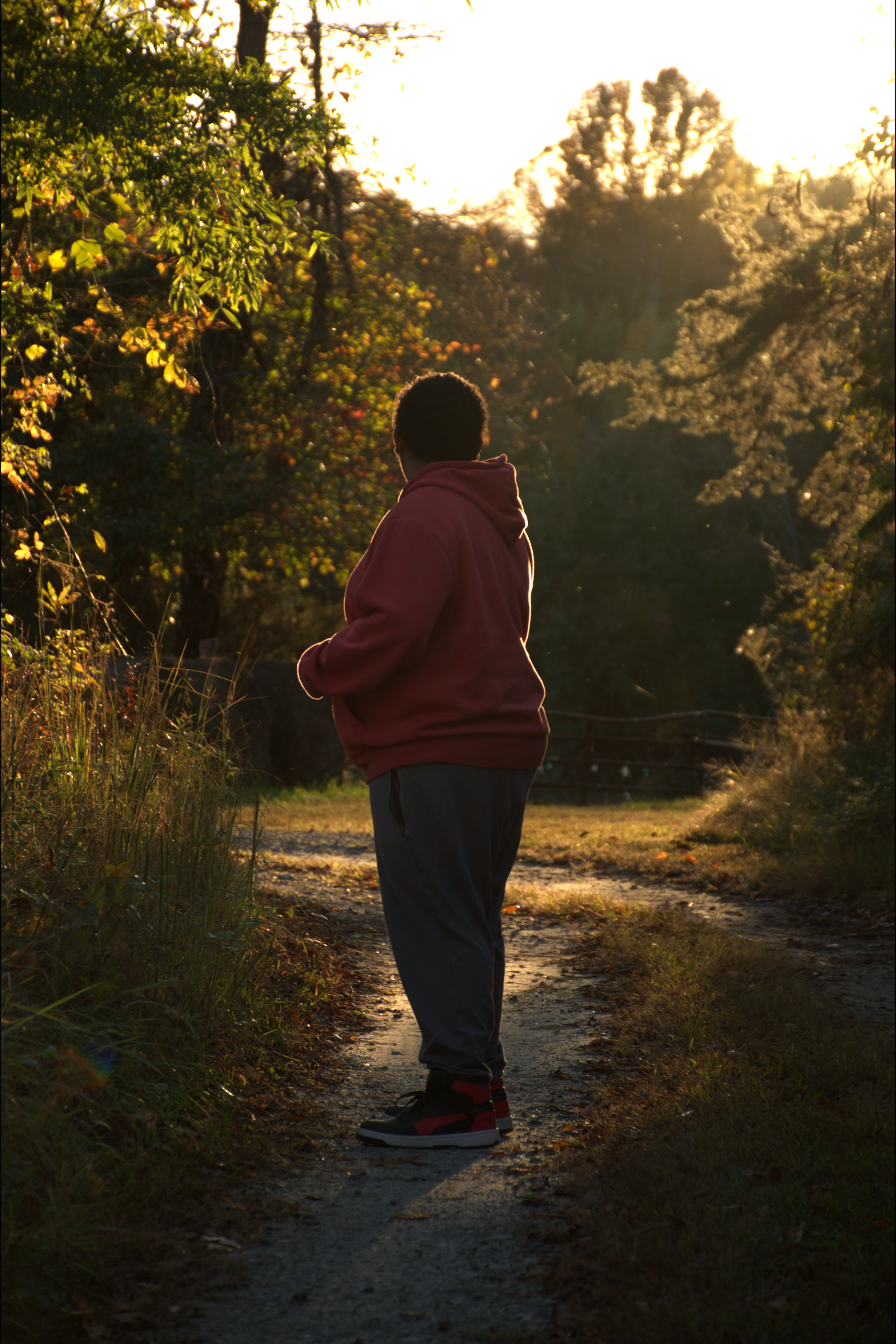 A person standing on a dirt path surrounded by trees and grass at sunset, wearing a red hoodie and black pants.