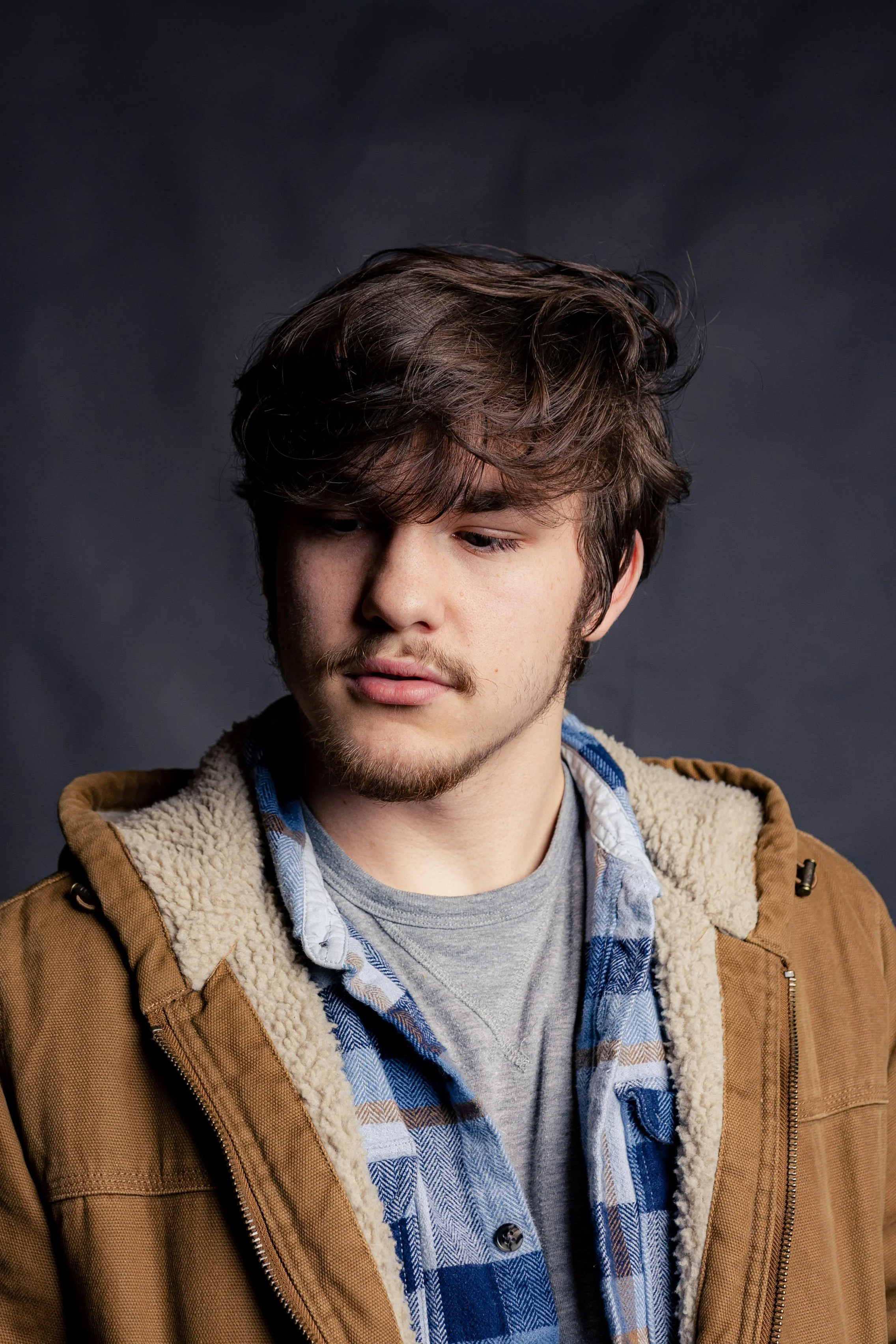 Portrait of a young man with brown hair and a mustache, wearing a brown jacket with a sherpa lining and a blue plaid shirt, against a dark background.