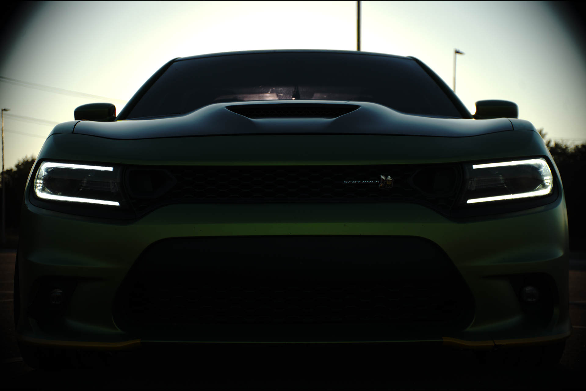 Front view of a dark-colored Dodge car with illuminated headlights, parked outdoors at dusk or dawn.