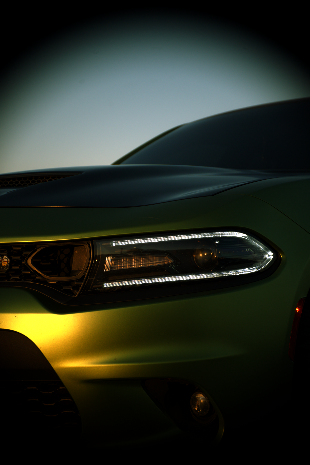 Close-up of a green car's front, showing the headlight and part of the hood at dusk or dawn.