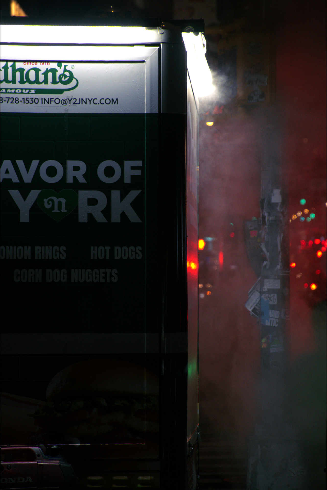 A food truck with a sign advertising onion rings, hot dogs, and corn dog nuggets. The truck is smoking in a city at night with visible traffic lights and street signs.