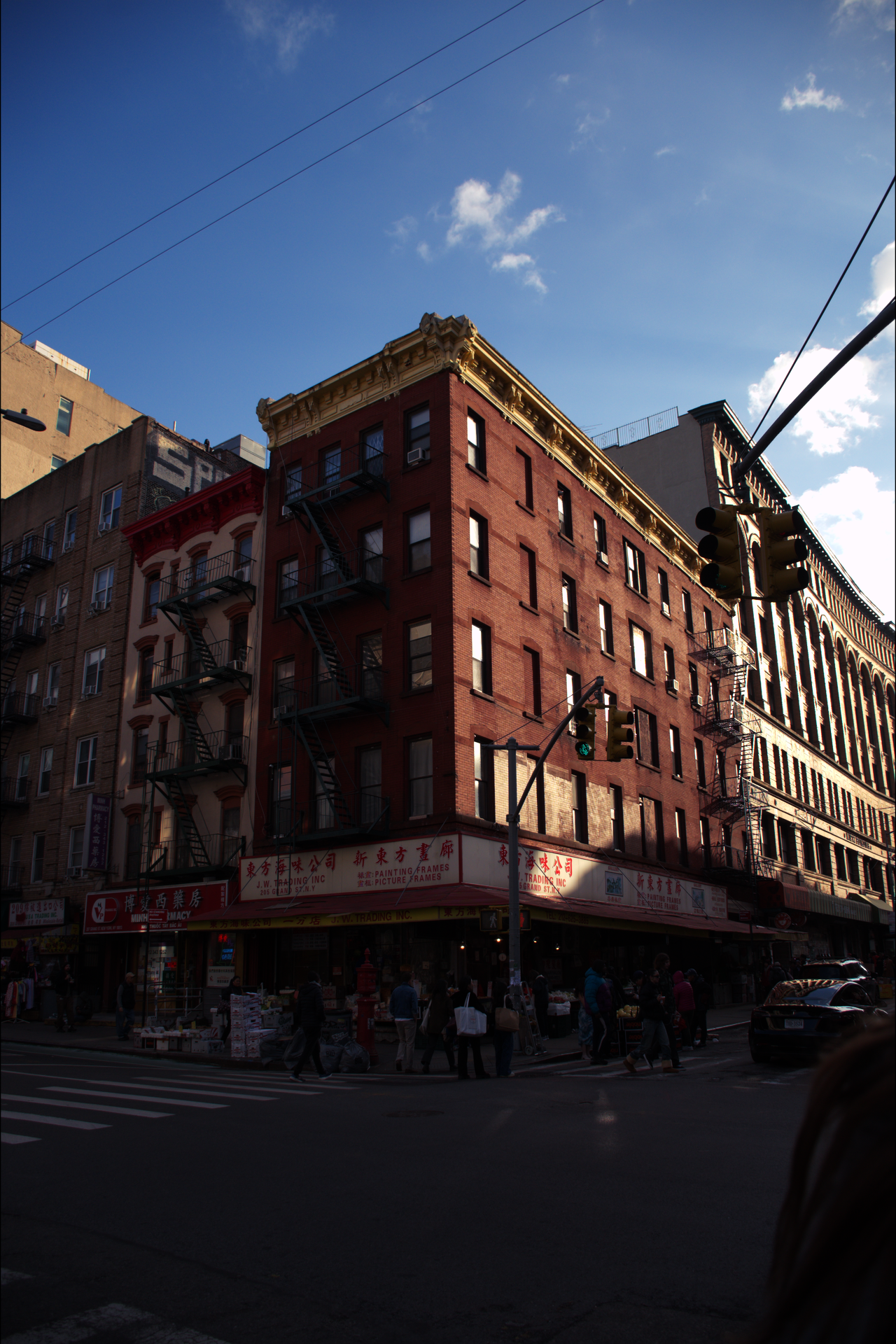 A city street scene featuring a corner building with a red brick facade, fire escape stairs, and a ground-floor store with Chinese signage, against a bright blue sky with white clouds.