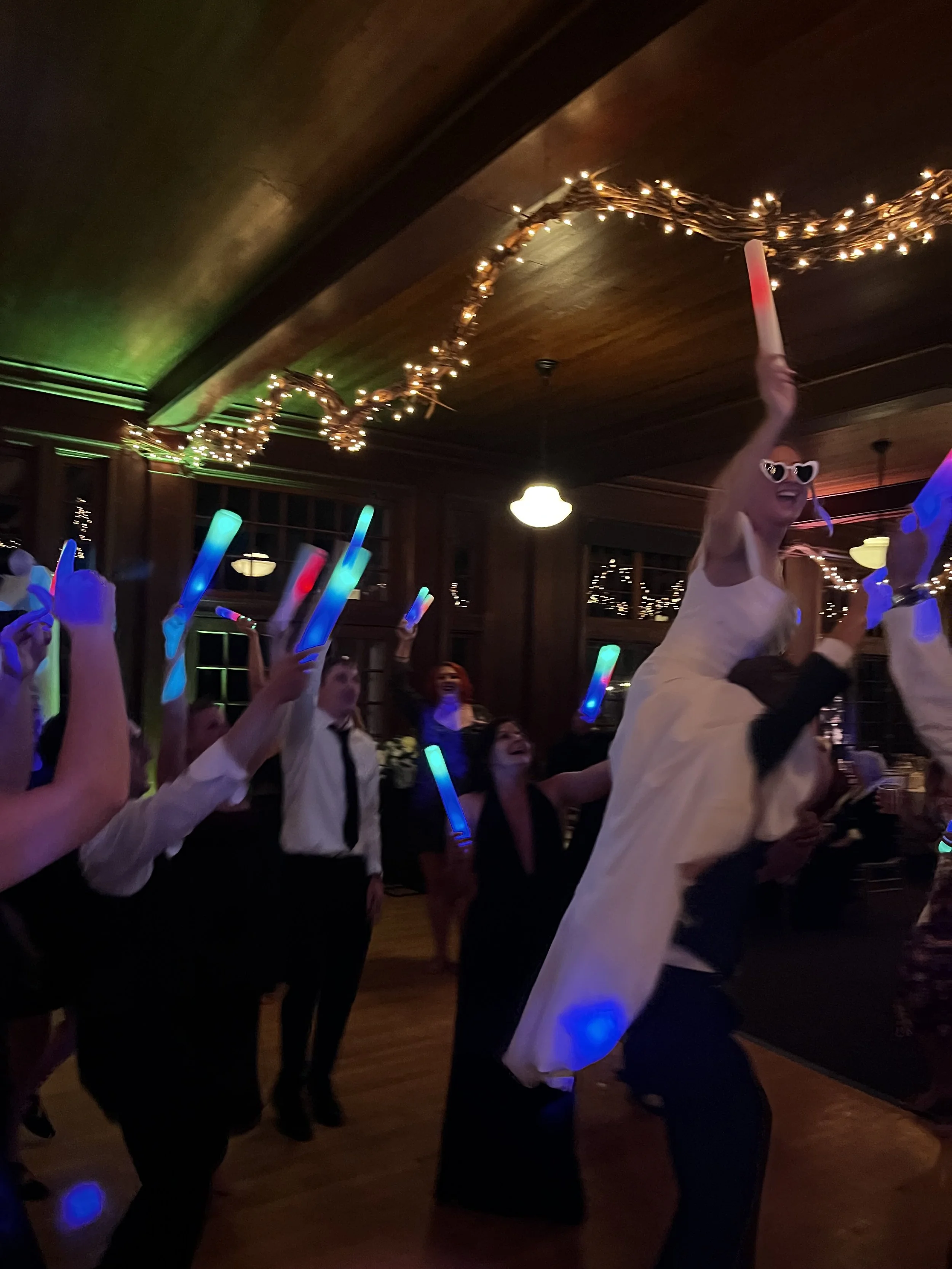 People dancing at a celebration, holding glow sticks, with a woman on shoulders wearing sunglasses and a white dress, in a decorated indoor venue with string lights.