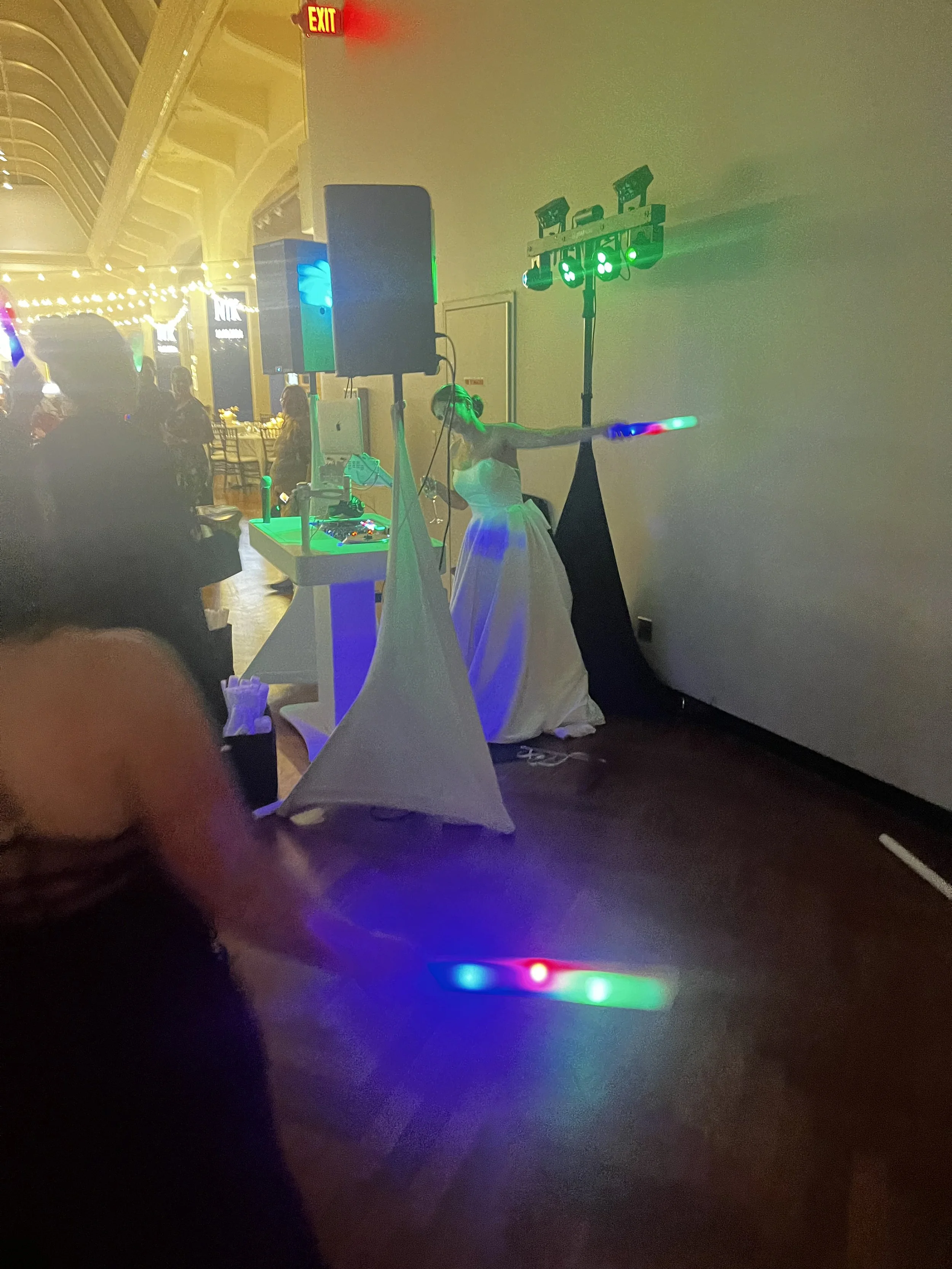 A woman in a white gown dancing with colorful LED light sticks in a party or wedding reception setting with dim lighting and other guests in the background.