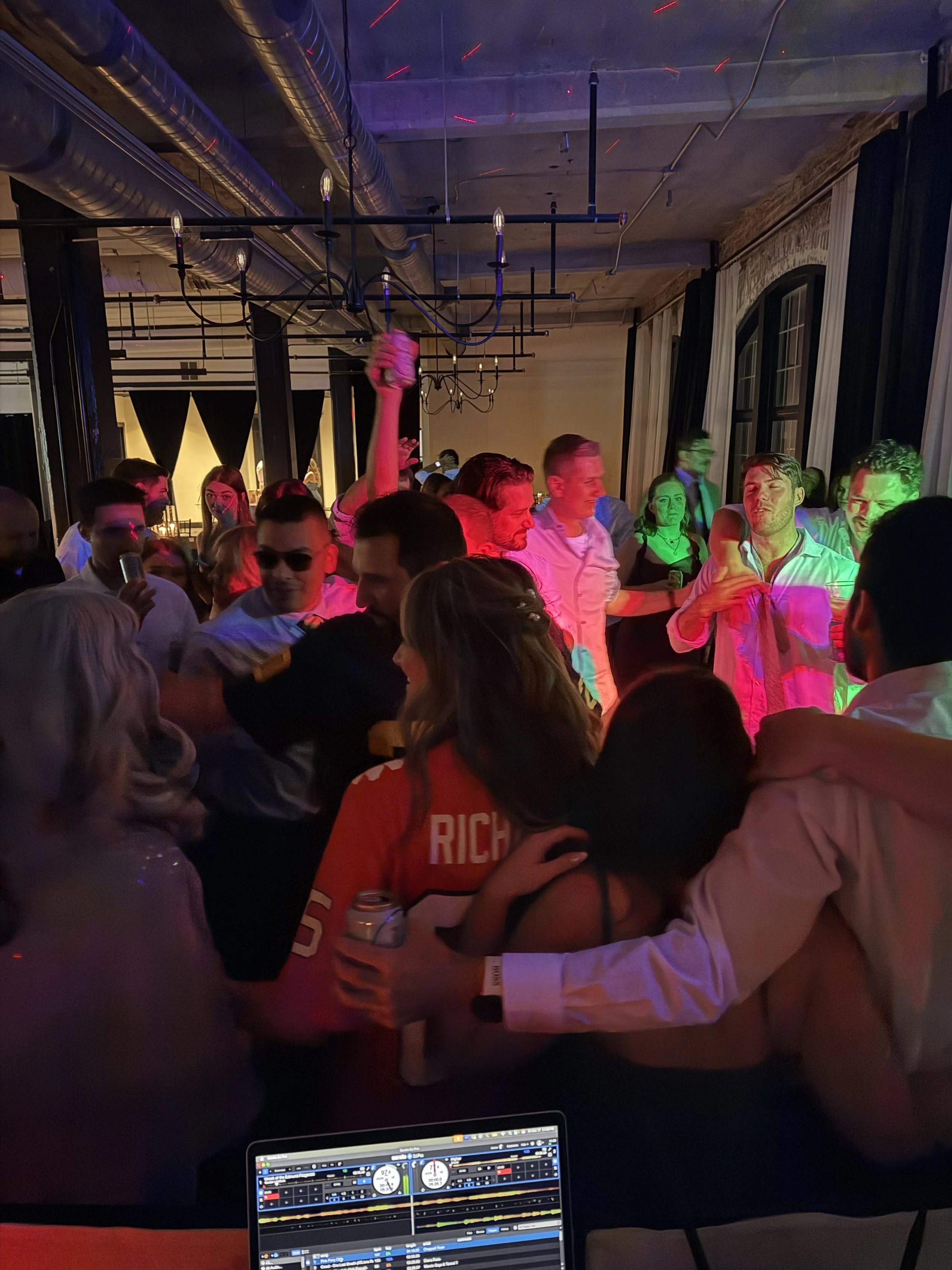 People dancing and socializing at a party or nightclub with colorful lighting and a DJ setup visible in the foreground.
