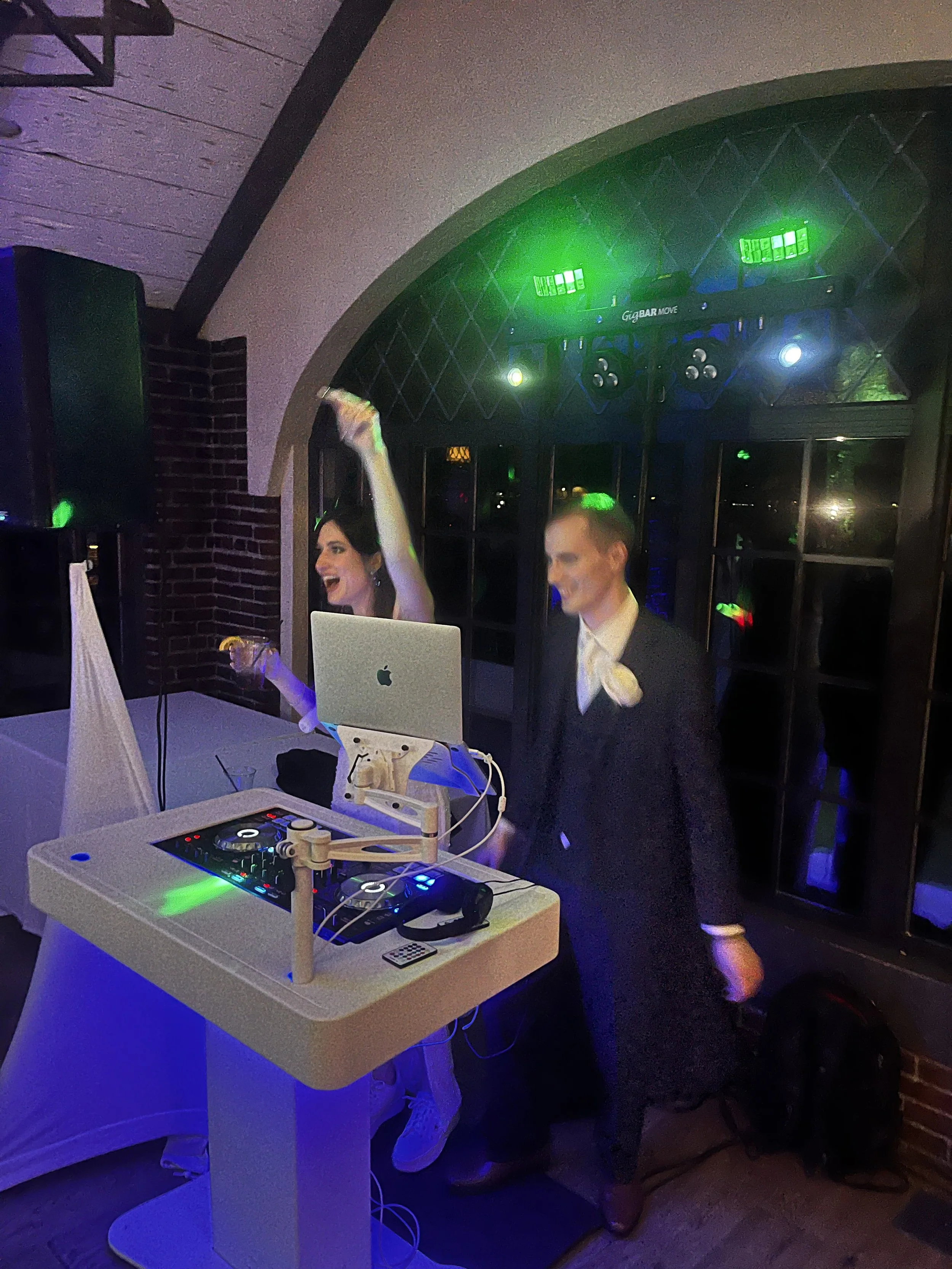 A woman and a man dancing and having fun at a party with a DJ setup and colorful lighting.