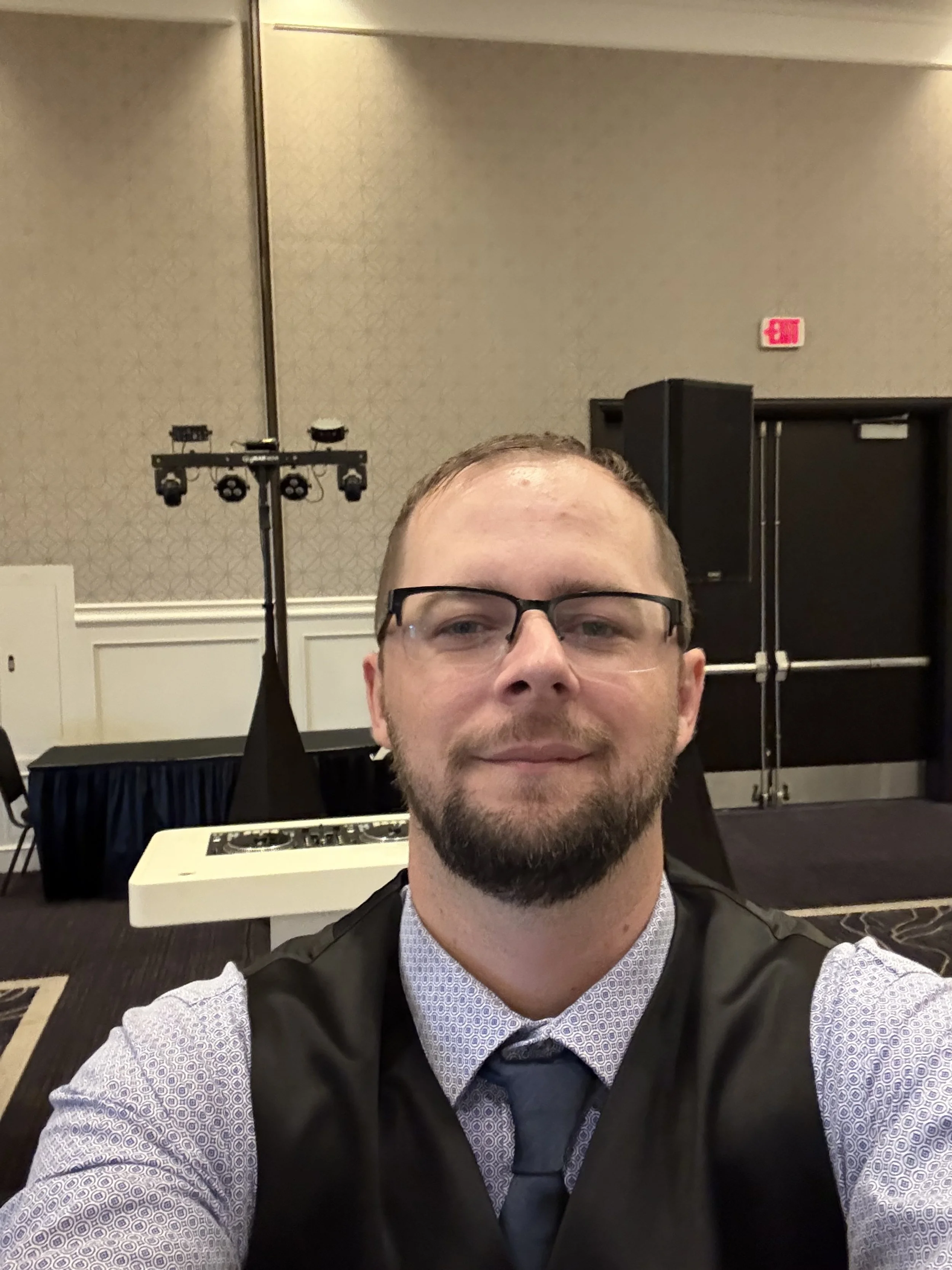 A man wearing glasses, a collared shirt, tie, and vest taking a selfie in a conference or event room with stage equipment and a black curtain in the background.
