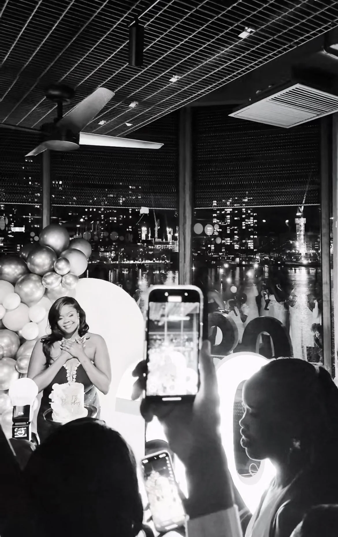 A woman celebrating her birthday at a party with a cake, surrounded by balloons and friends taking pictures, in a modern venue with city lights visible through large windows.