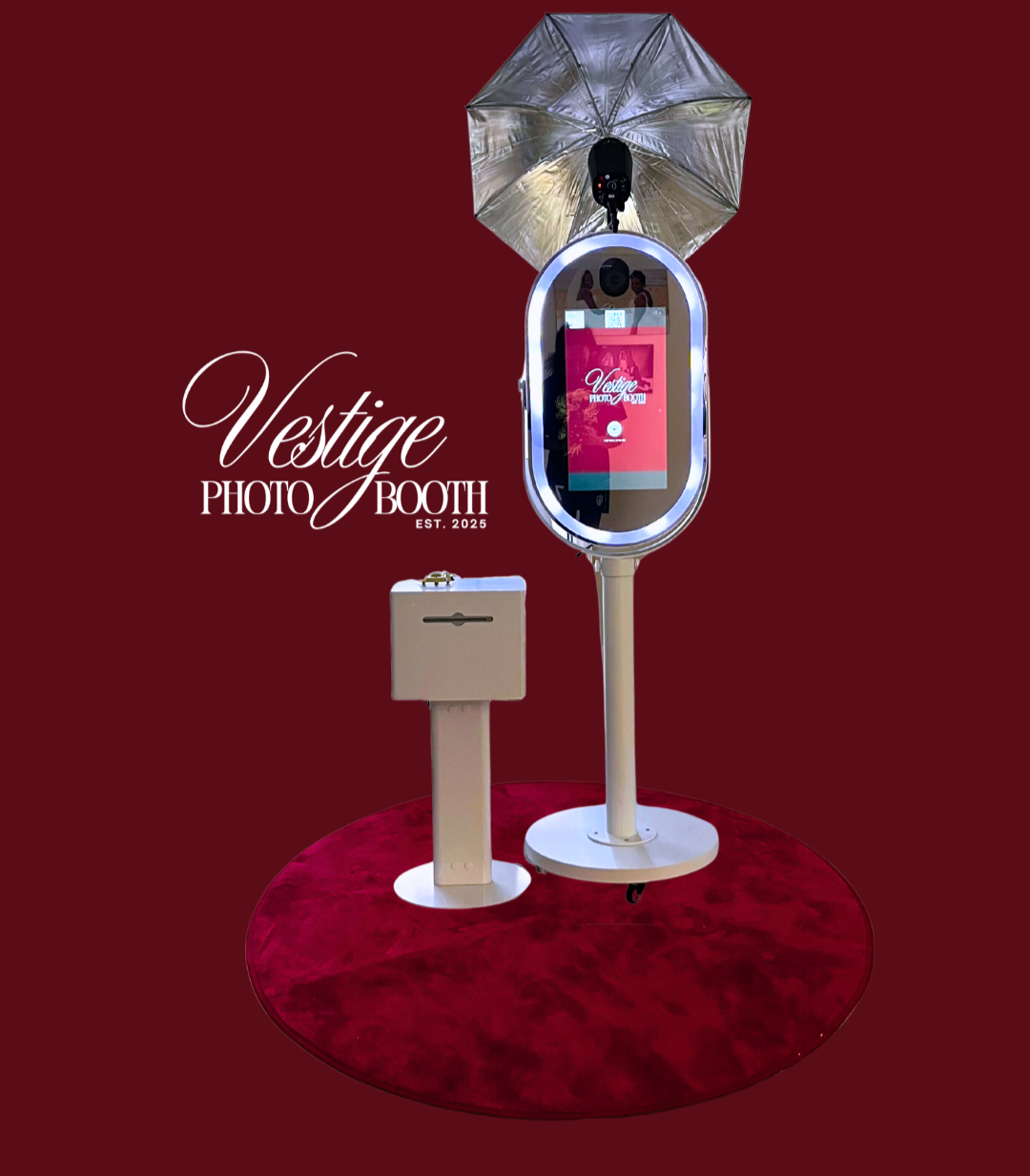 A modern photo booth setup on a red carpet with a red backdrop. The booth has a sleek, oval-shaped design with a digital screen, a camera, and an umbrella for lighting. Next to it is a small beige box on a stand, possibly for accessories or props. The logo 'Vestige Photo Booth' and establishment year 2025 are displayed on the backdrop.