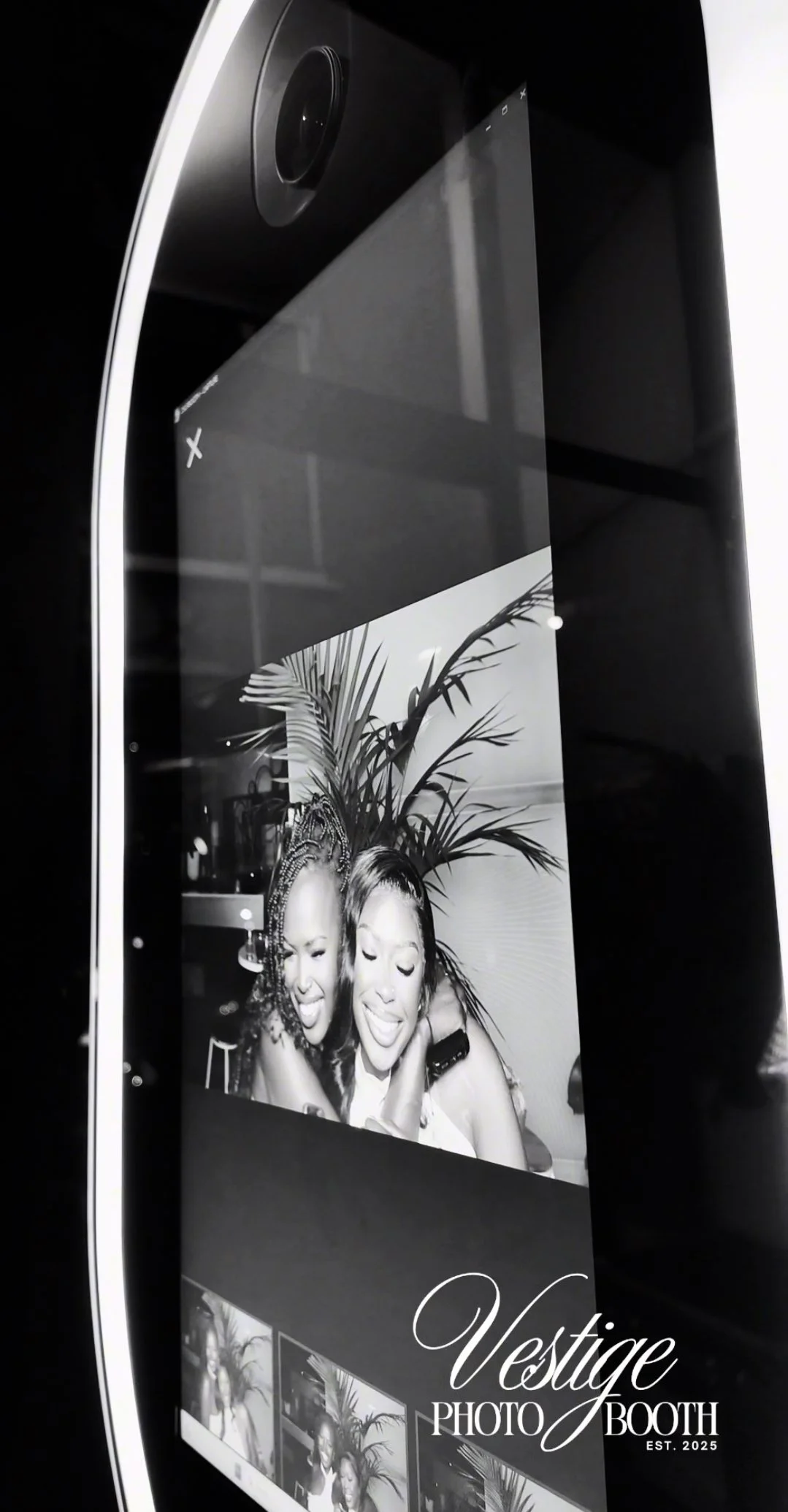 A digital photo booth screen displaying a black-and-white photo of two women smiling and hugging, with a palm plant in the background. The photo booth branding 'Vesige Photo Booth Est. 2025' is visible at the bottom.