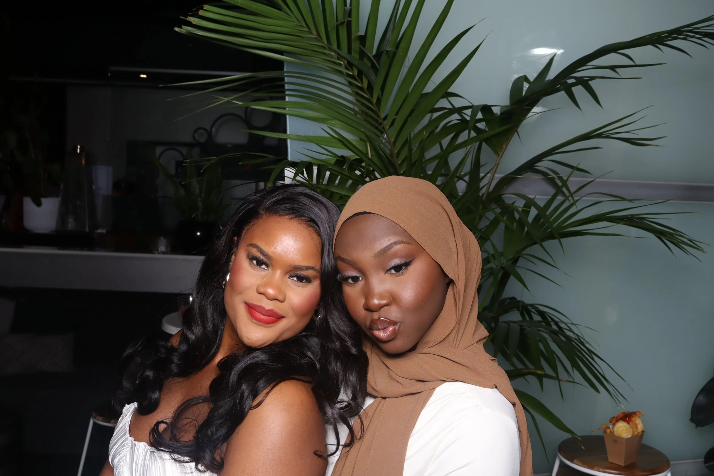 Two women posing together indoors, one with black wavy hair and red lipstick, the other wearing a tan hijab, with a large green plant in the background and a table with a small plate of food in the corner.