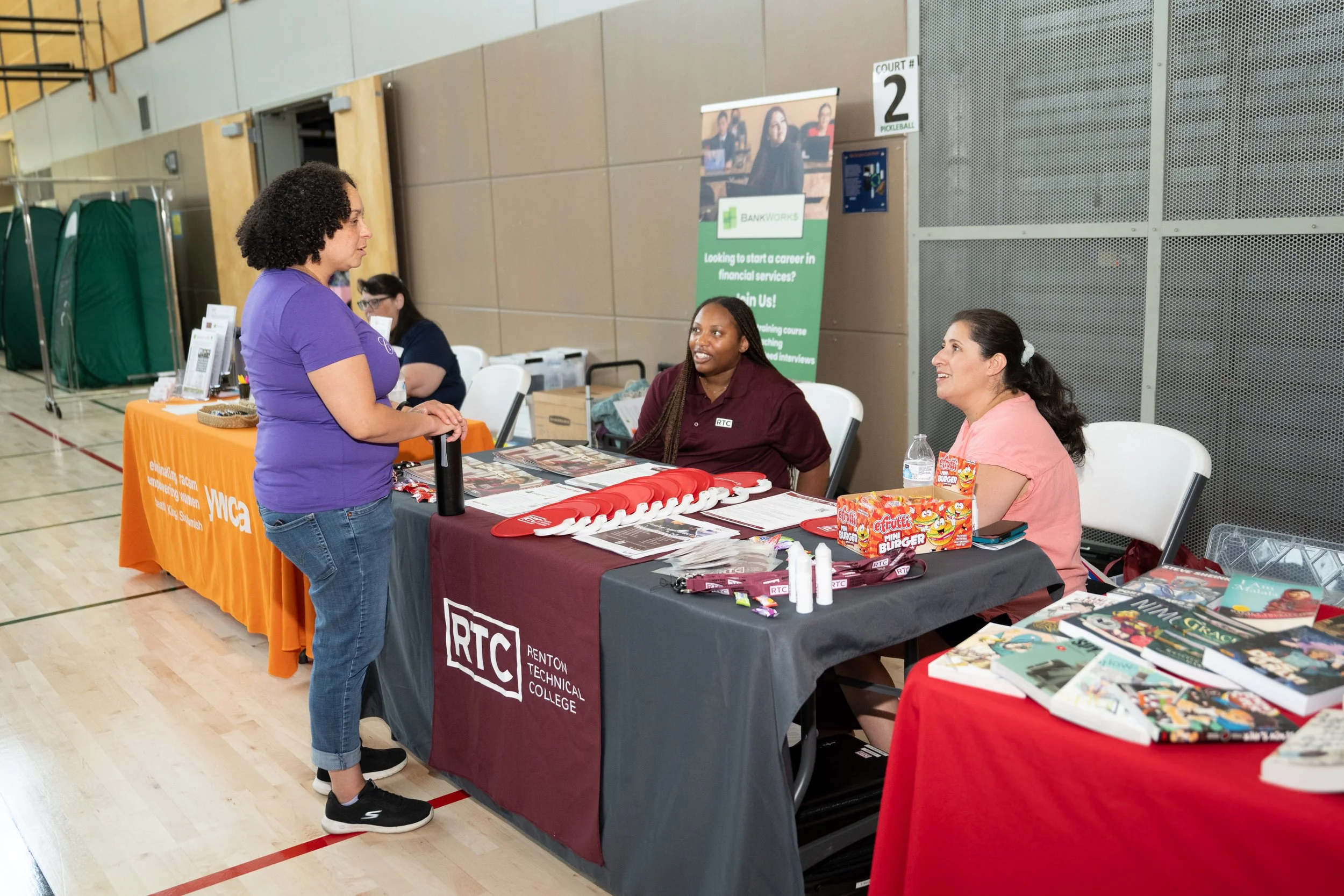 A woman in a purple shirt and jeans is talking to two women sitting behind a table at a career fair. The table has promotional materials from Renton Technical College and RTC, with various pamphlets, lanyards, and small items. The background includes another booth with an orange tablecloth for YWCA and a promotional banner for BankWorks.