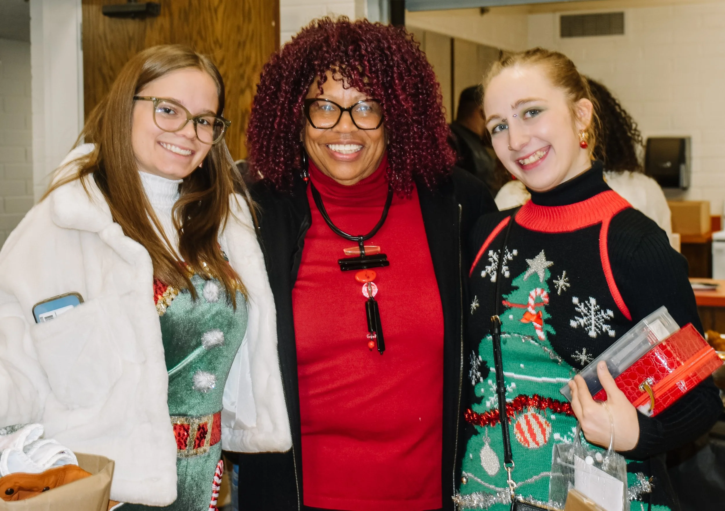 Three women smiling at a Christmas event, wearing festive sweaters, with the woman in the center wearing glasses and a red top.