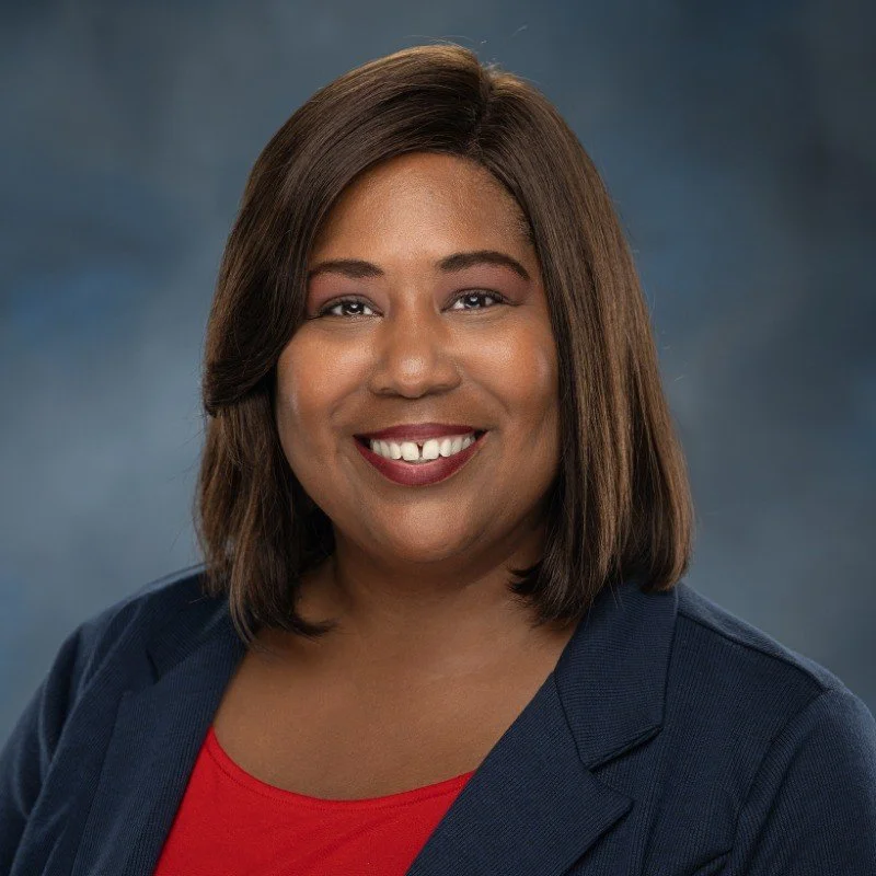 A smiling woman with shoulder-length brown hair wearing a navy blazer and red top against a dark blue background.