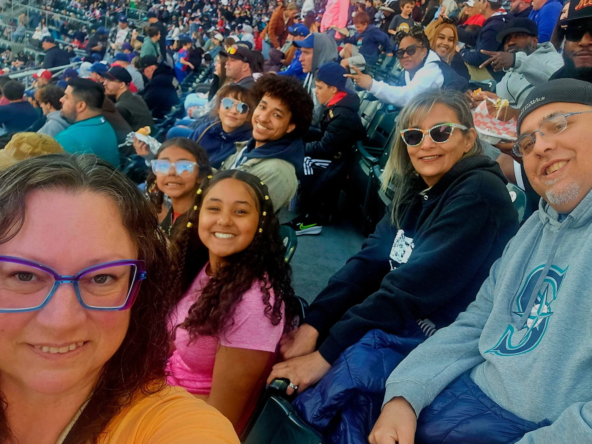 A group of smiling people at a sporting event sitting on stadium seats, wearing casual clothing and sunglasses, with many other spectators in the background.
