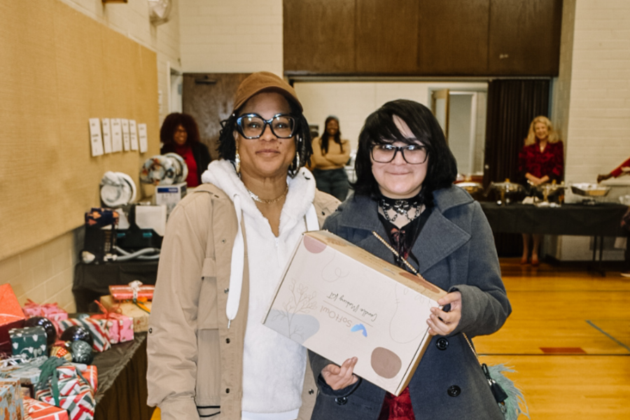 Two women standing indoors at a celebration event. One is wearing a tan jacket and hat, glasses, and earrings, while the other is in a gray coat with black hair and glasses, holding a large gift box. Several people and tables with presents are visible in the background.