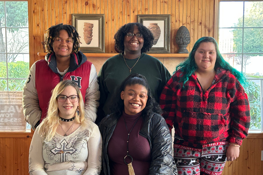 Five women posing for a group photo indoors in front of a wooden wall with framed artwork and large windows showing trees outside. They are smiling and dressed casually.