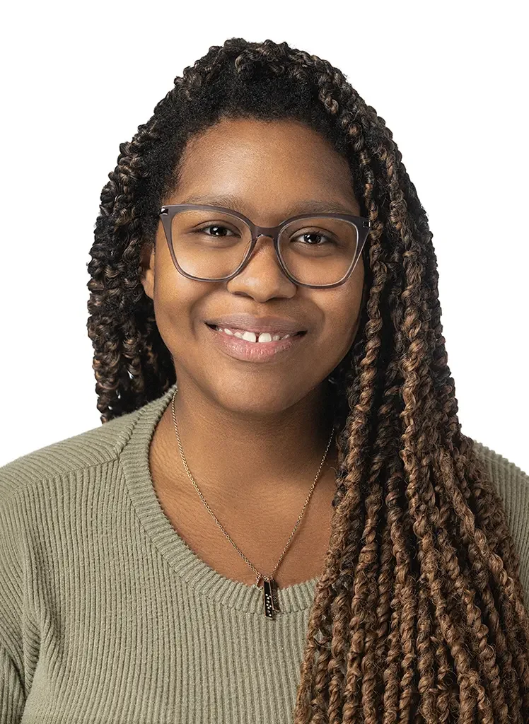 Portrait of a smiling young woman with glasses and long curly hair, wearing a green sweater and a necklace, against a white background.