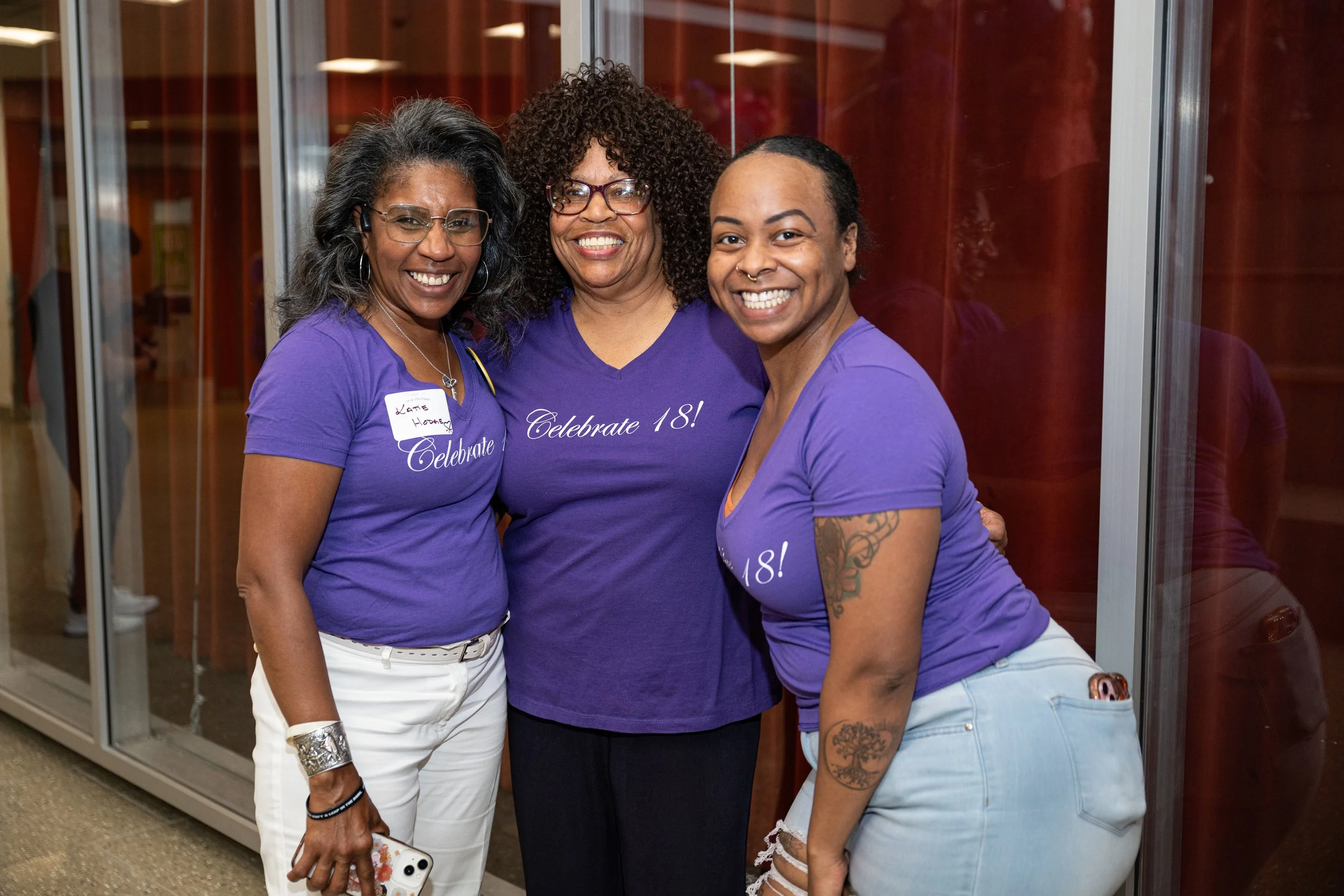 Three women wearing purple shirts celebrating, standing together and smiling at the camera in a room with glass doors. The shirts have 'Celebrate 18!' written on them.